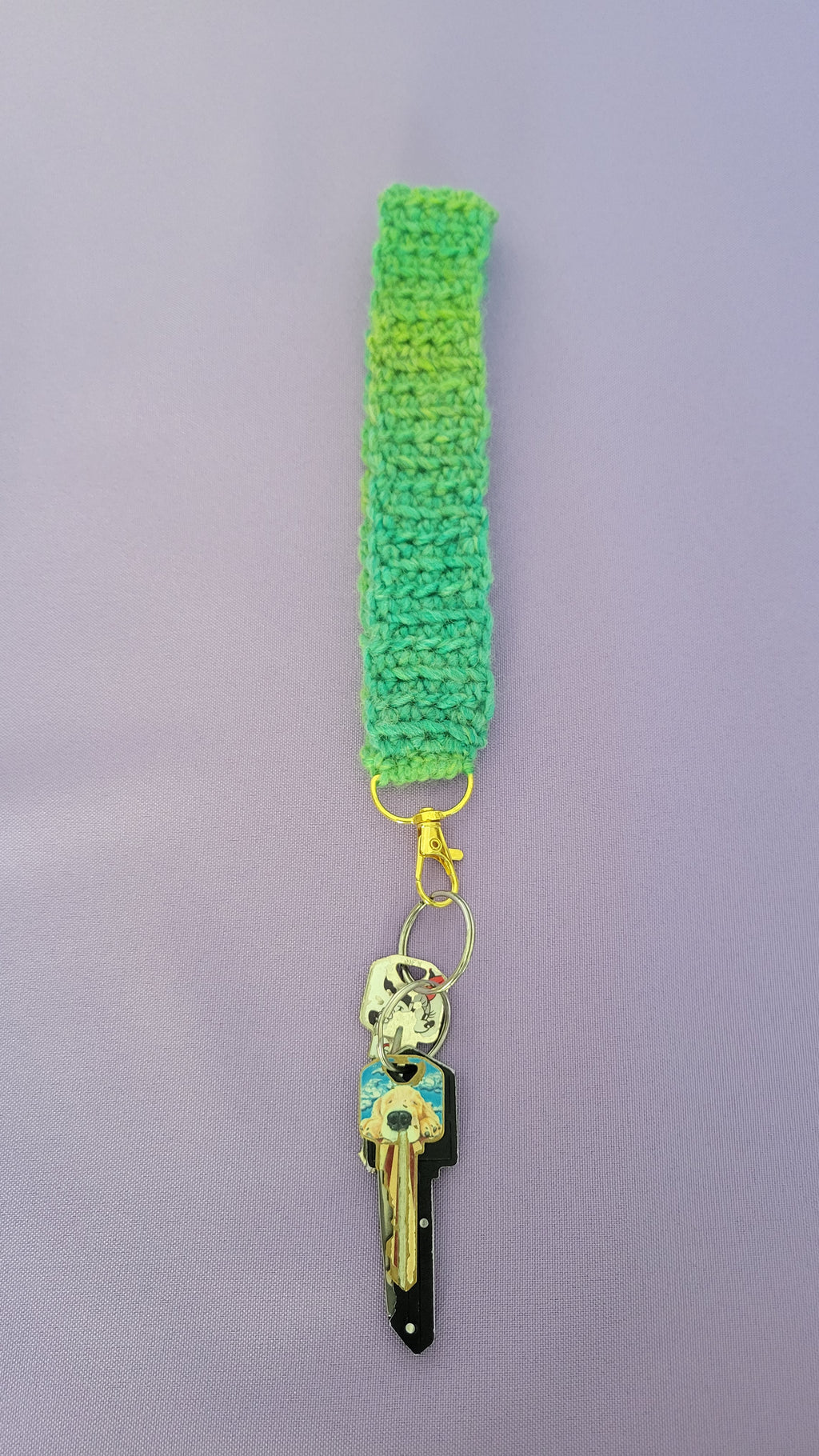 Green crochet key bracelet item with a key attached,against a purple background. The size of the bracelet size is nine inches.