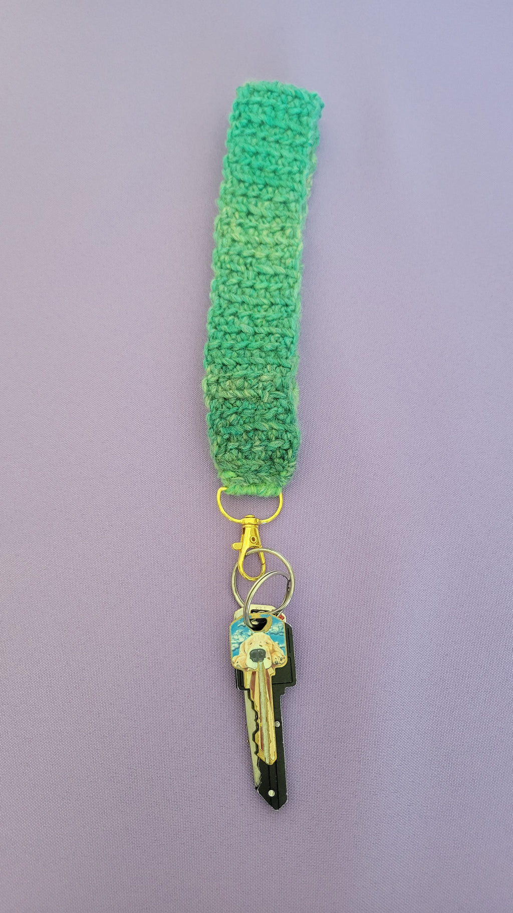 Green crochet key bracelet item with a key attached,against a purple background. The size of the bracelet is eight inches.
