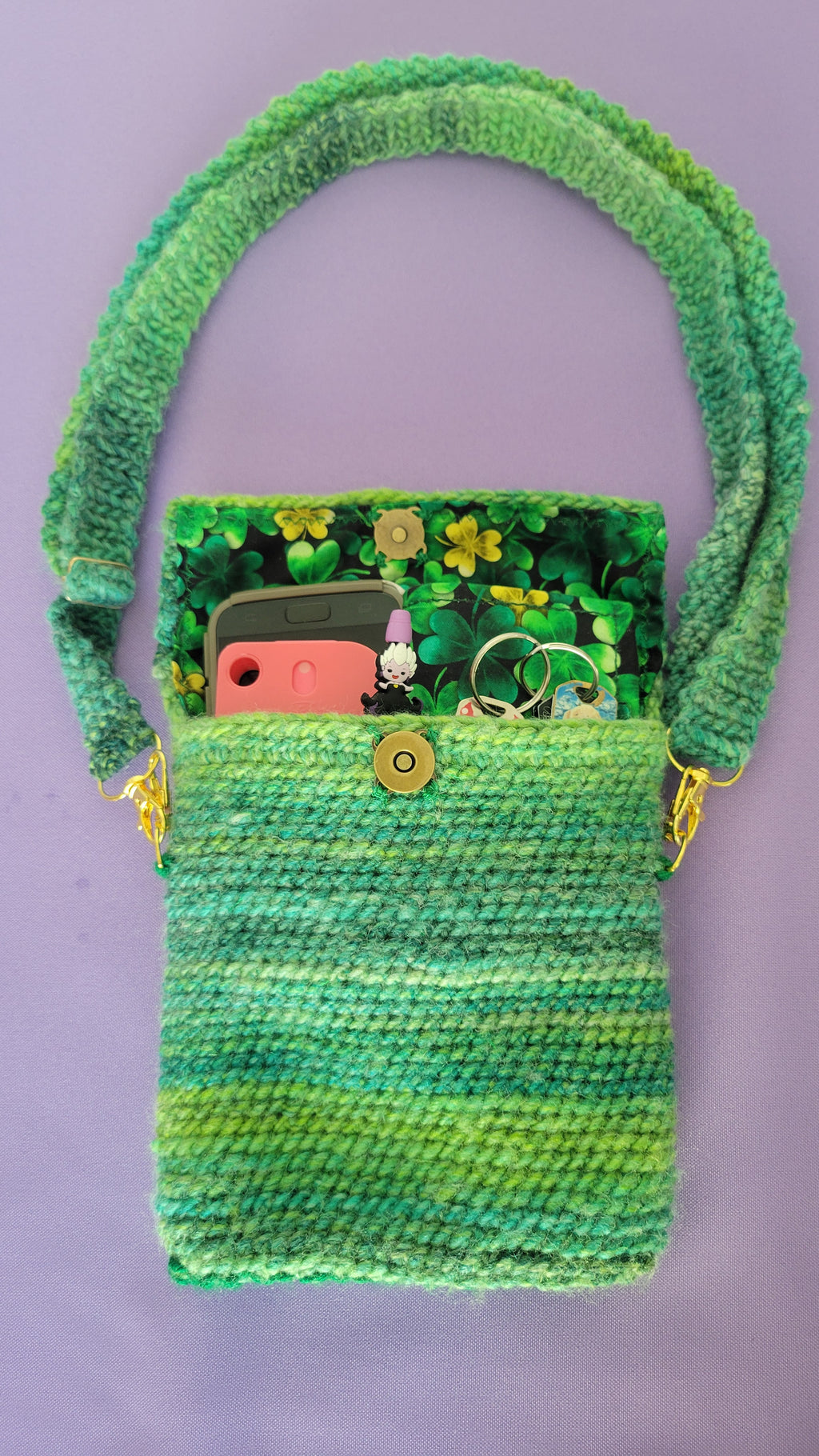 A crochet purse from the laying down angle. The strap is attach with gold accessories, the bronze magnetic closure is visible and crochet together by a multiple shades of green yarn against purple background. Inside, you can see the clover interfacing, and the items the purse can hold. There is a hand sanitizer, a pen, a touch phone, keys and wallet.