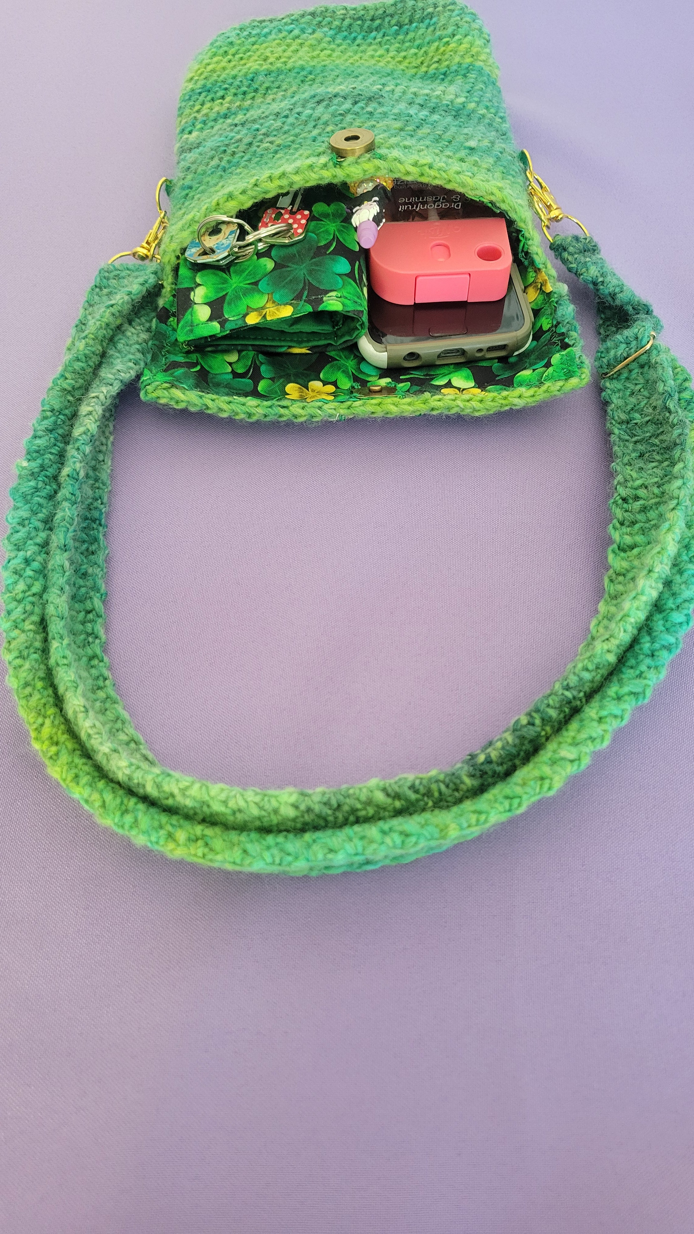 A crochet purse from the laying down position from a front angle. The strap is attach with gold accessories, the bronze magnetic closure is visible and crochet together by a multiple shades of green yarn against purple background. Inside, you can see the clover interfacing, and the items the purse can hold. There is a hand sanitizer, a pen, a touch phone, keys and wallet.