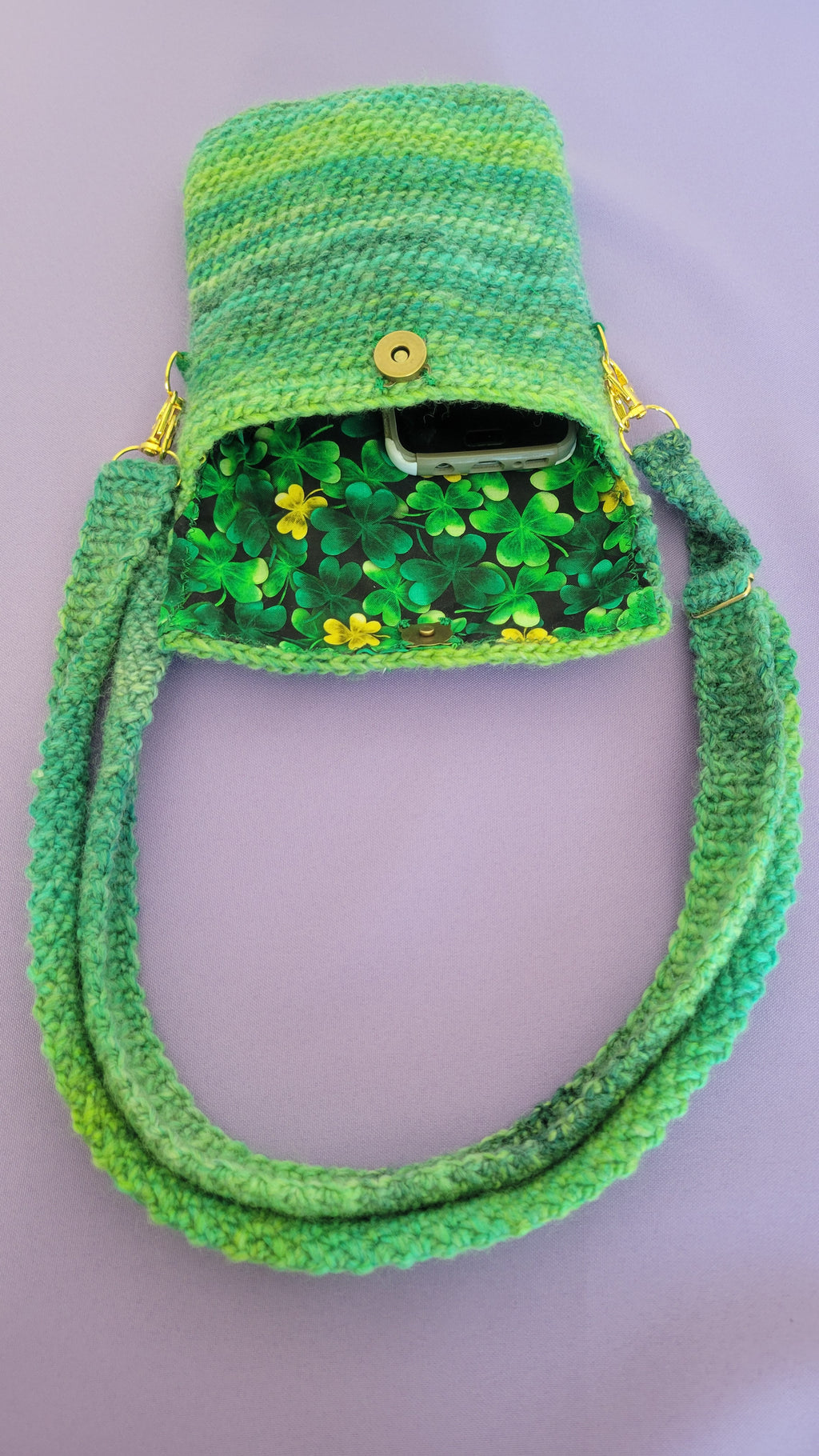 A crochet purse from the inside angle. Showcasing the clover design and the amount of space the purse can hold. The strap is attach with gold accessories, the bronze magnetic closure is visible and crochet together by a multiple shades of green yarn against purple background.