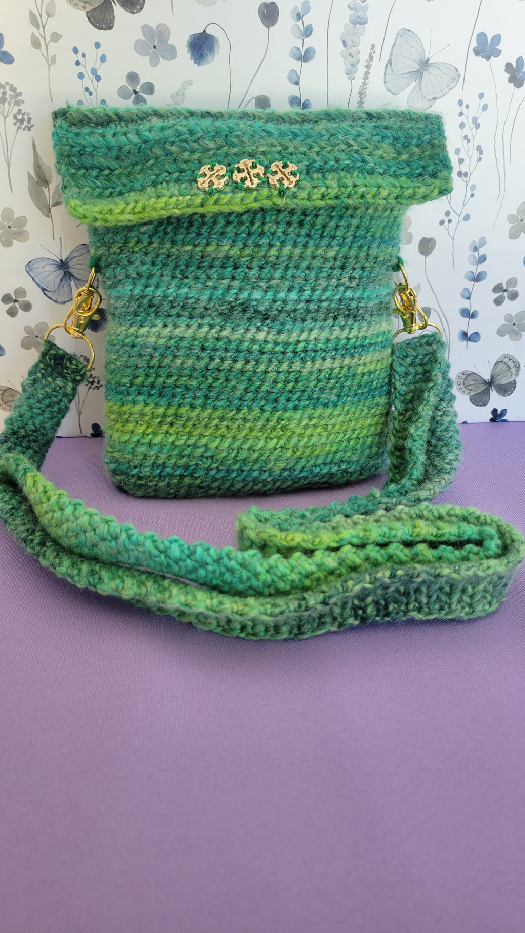 A crochet purse from the front angle. Showcasing three gold clovers.  The strap is attach with gold accessories, and crochet together by a multiple shades of green yarn against purple background.