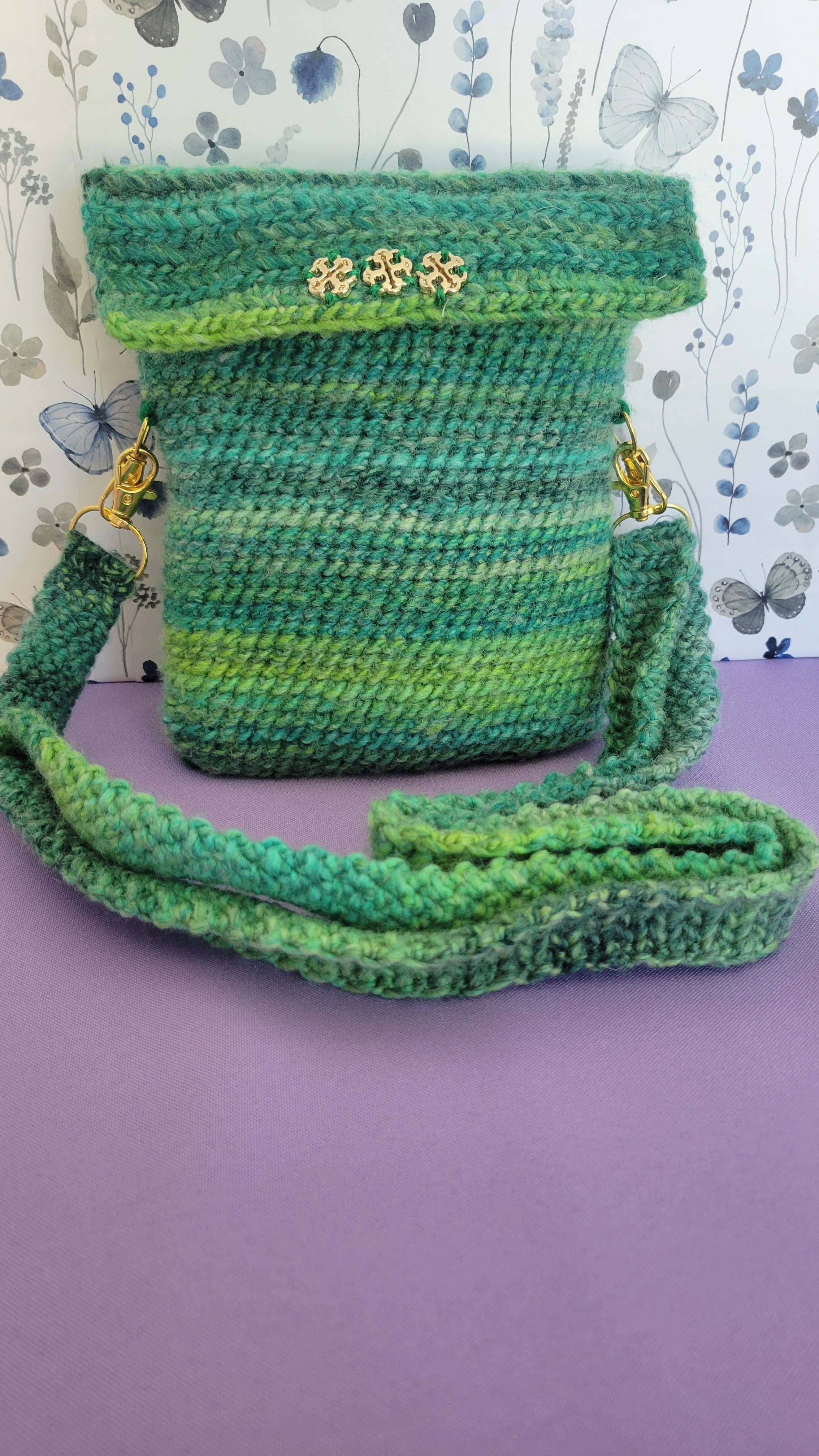 A crochet purse from the front angle. Showcasing three gold clovers.  The strap is attach with gold accessories, and crochet together by a multiple shades of green yarn against purple background.