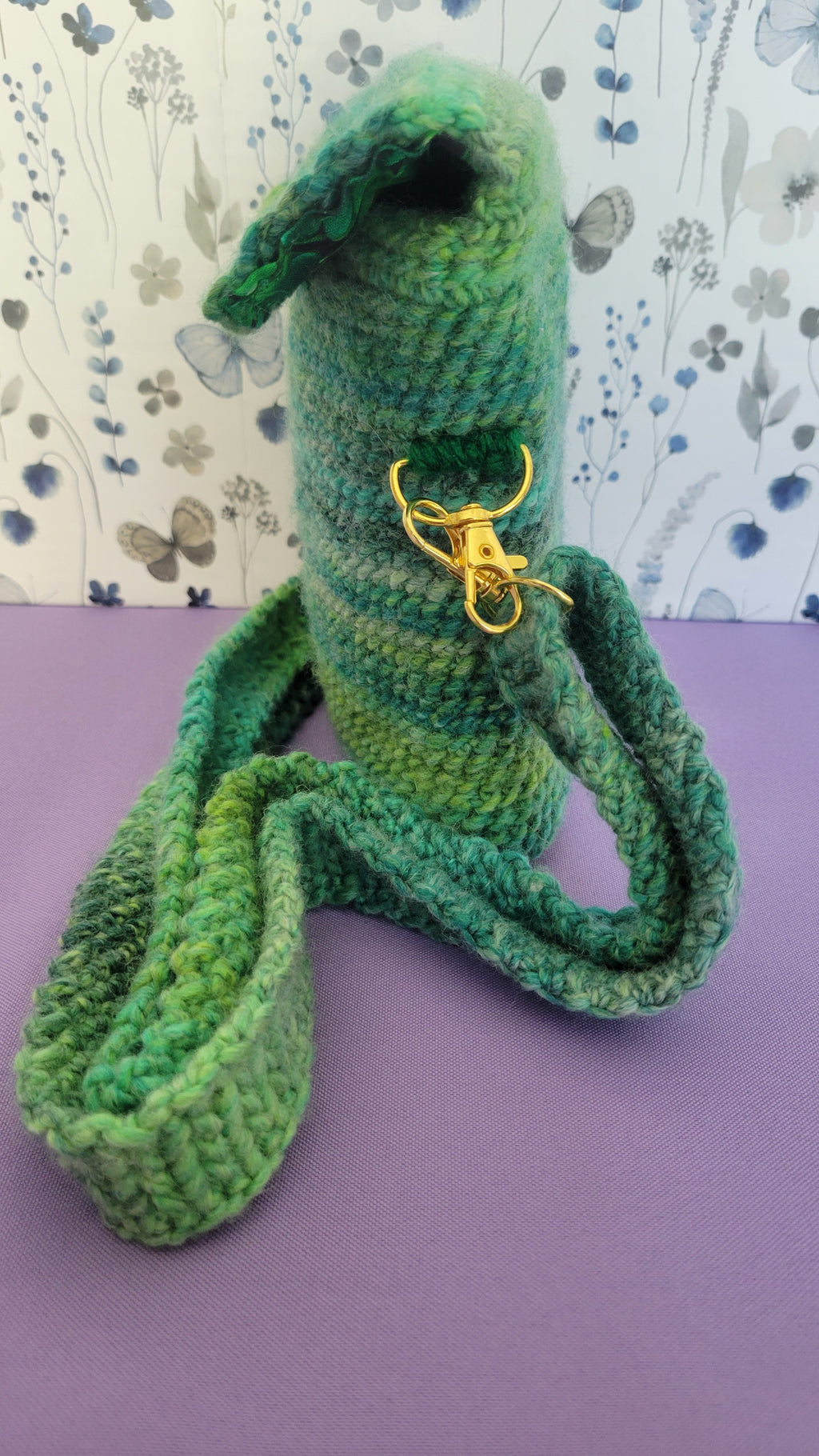 A crochet purse from the side angle. The strap is attach with gold accessories, and crochet together by a multiple shades of green yarn against purple background.