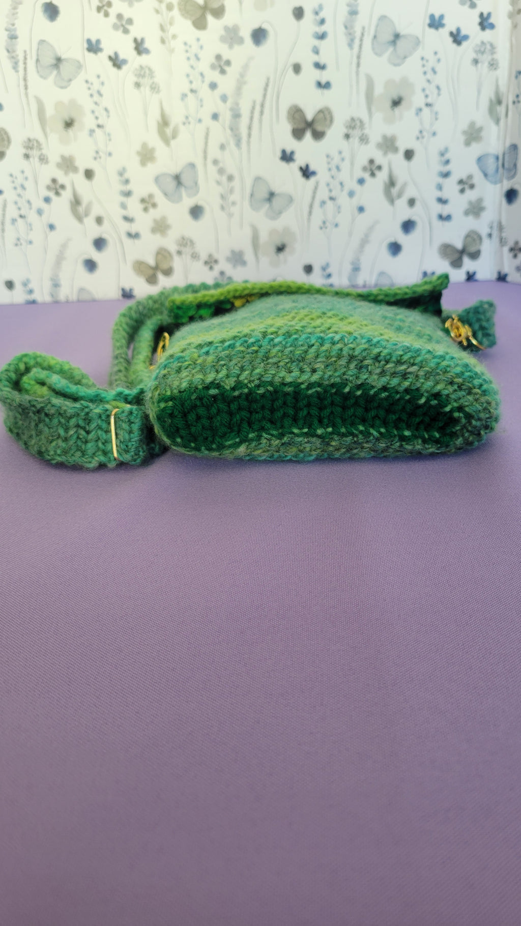 A crochet purse from a bottom angle. The strap is attach with gold accessories, and crochet together by a multiple shades of green yarn against purple background.