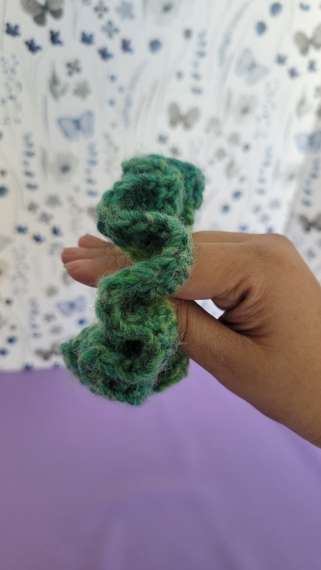 Green crochet scrunchy in hand. Surrounded by blue and grey butterflies on a white and purple background