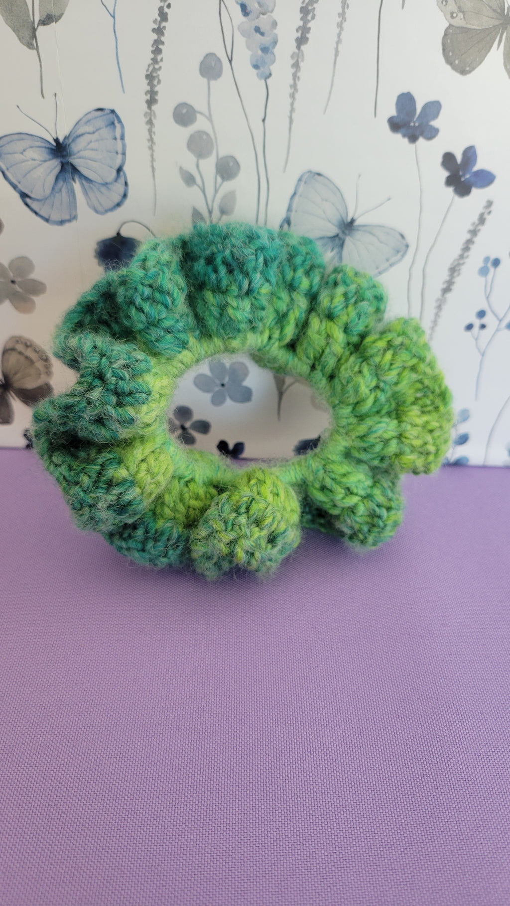 Green crocheted scrunchie with a small animal design on a purple and floral-patterned background