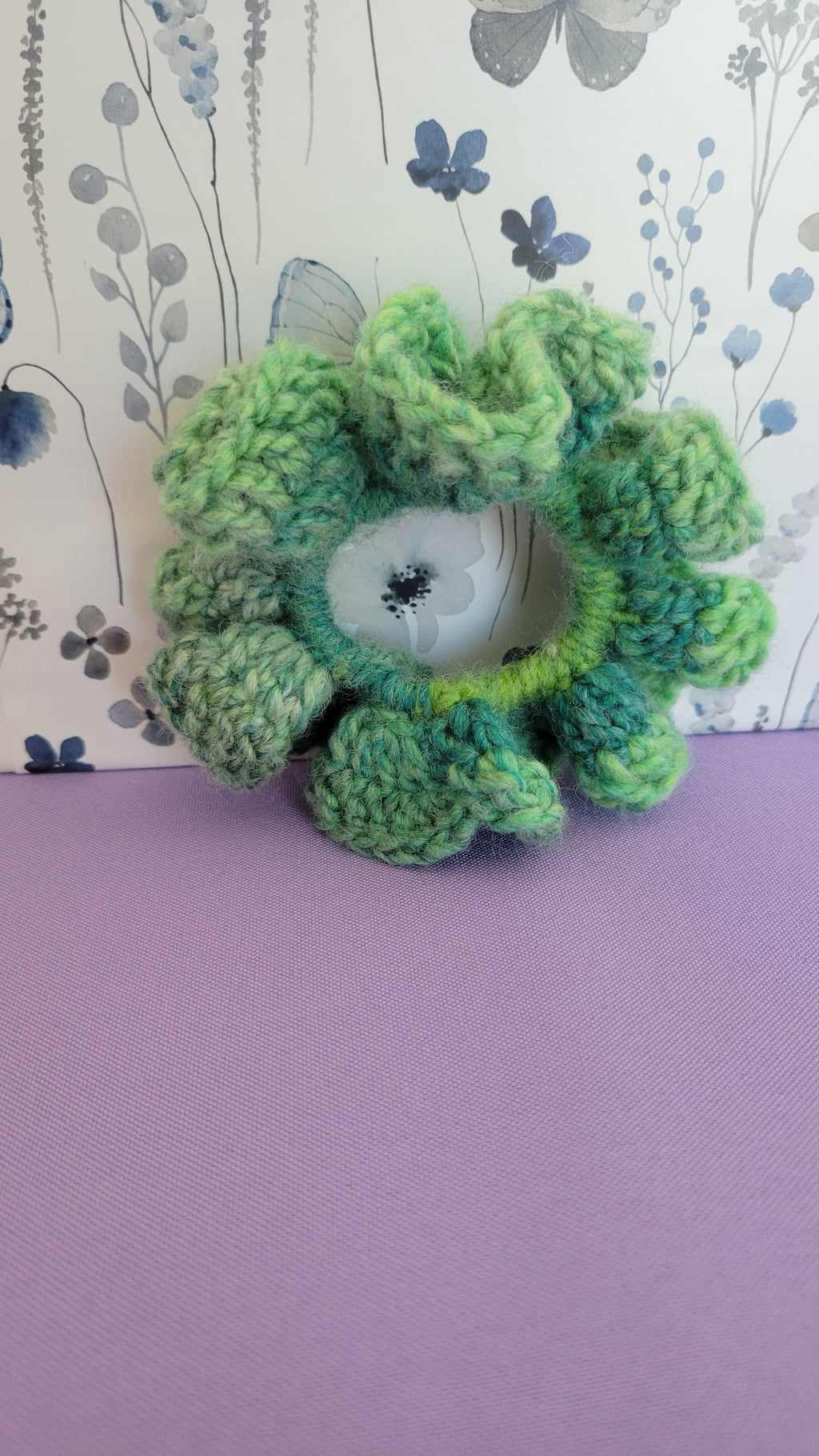 Green crochet scrunchy standing up. Surrounded by blue and grey butterflies on a white and purple background