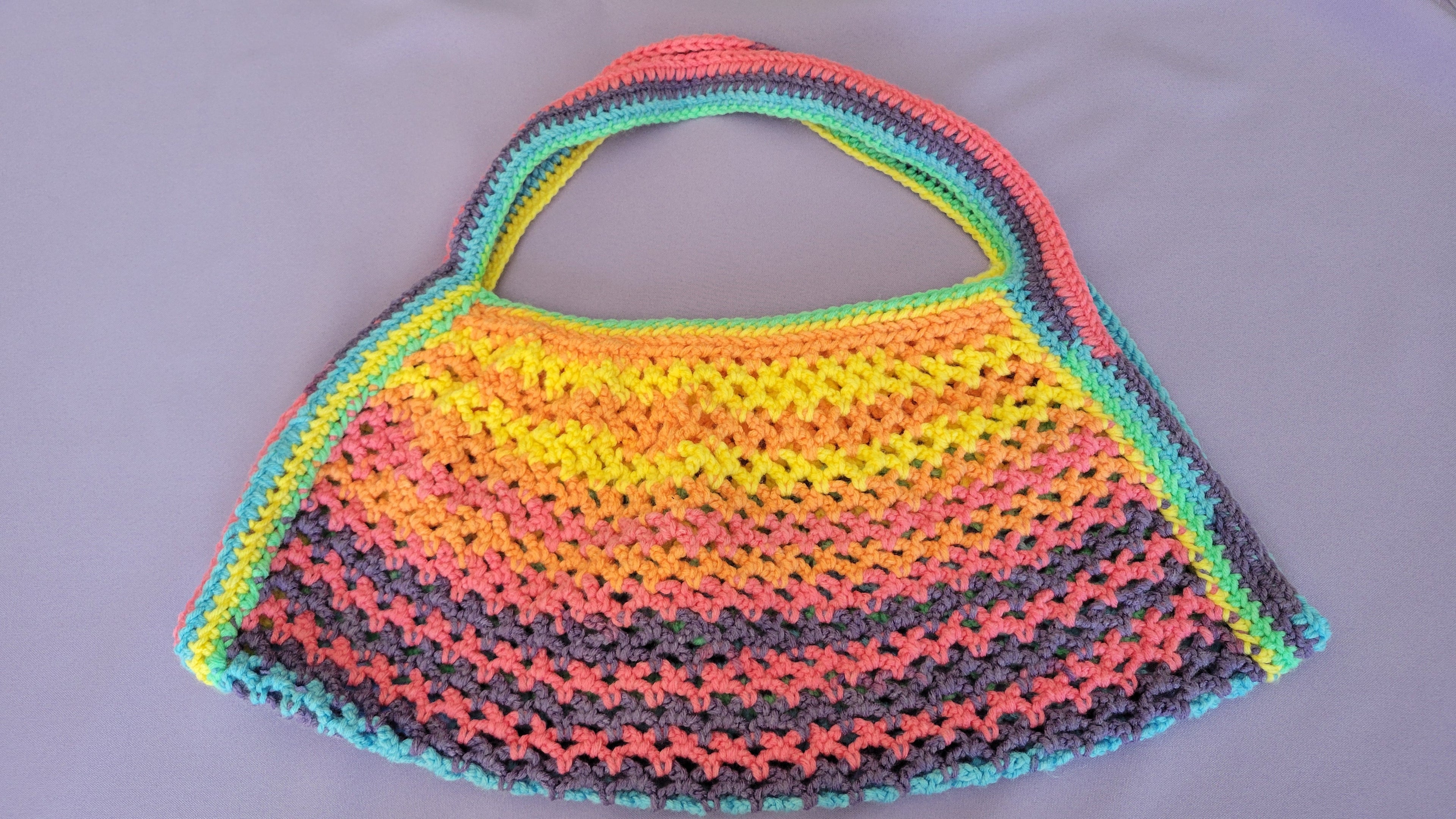 A handmade crochet beach bag with a wavy striped pattern in multiple colors including yellow, orange, pink, gray, and green, laid flat on a gray background.