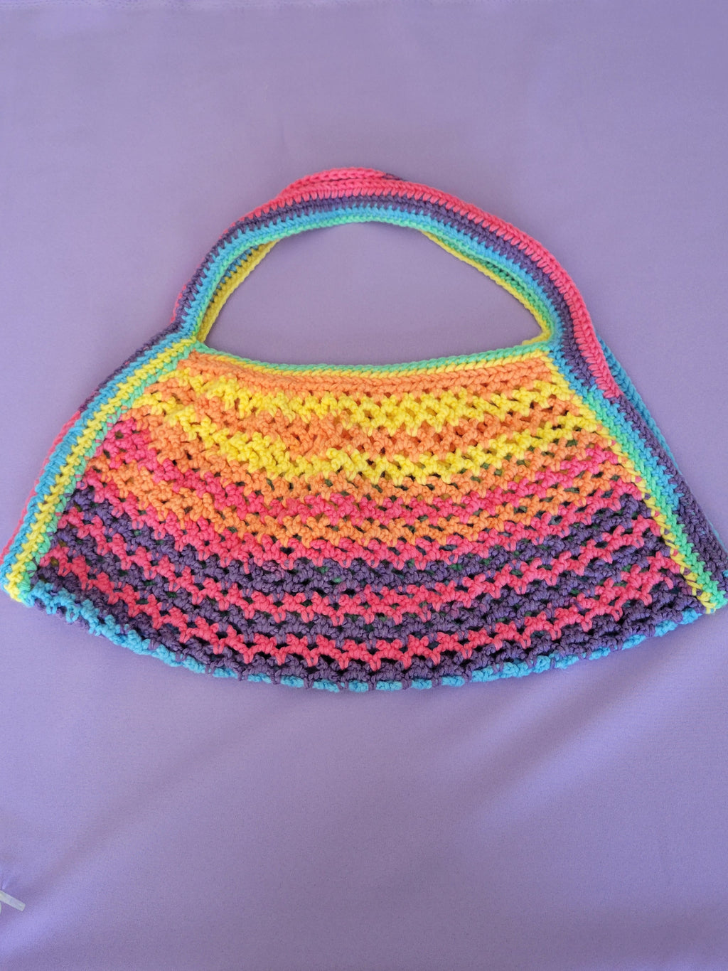 A handmade crocheted shoulder bag with a wide handle, featuring horizontal rainbow stripes of pink, orange, yellow, green, and blue on a light purple background.