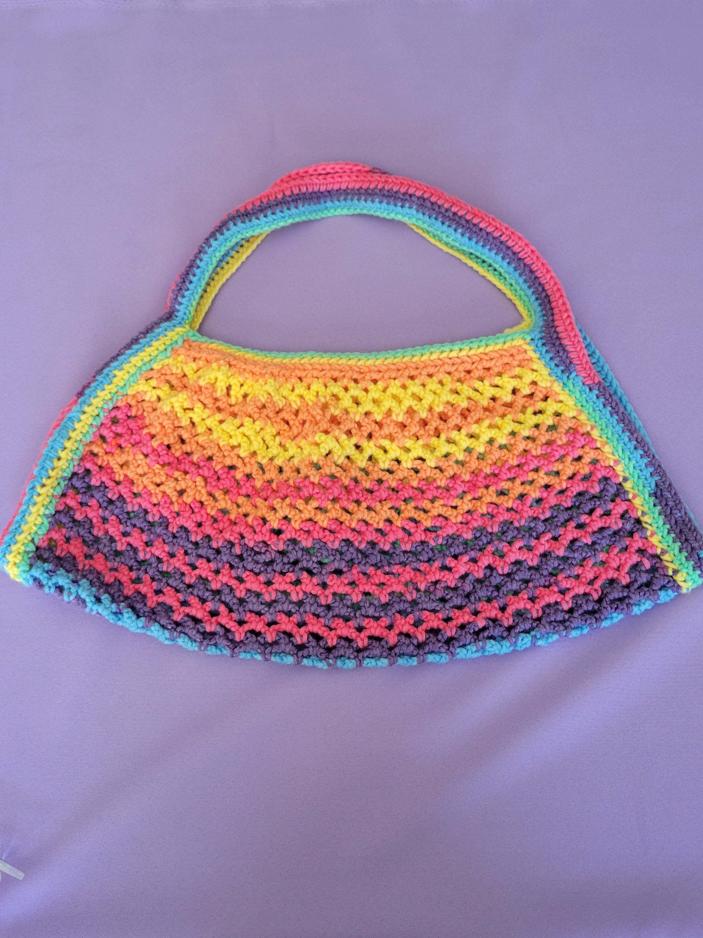 A handmade crocheted shoulder bag with a wide handle, featuring horizontal rainbow stripes of pink, orange, yellow, green, and blue on a light purple background.