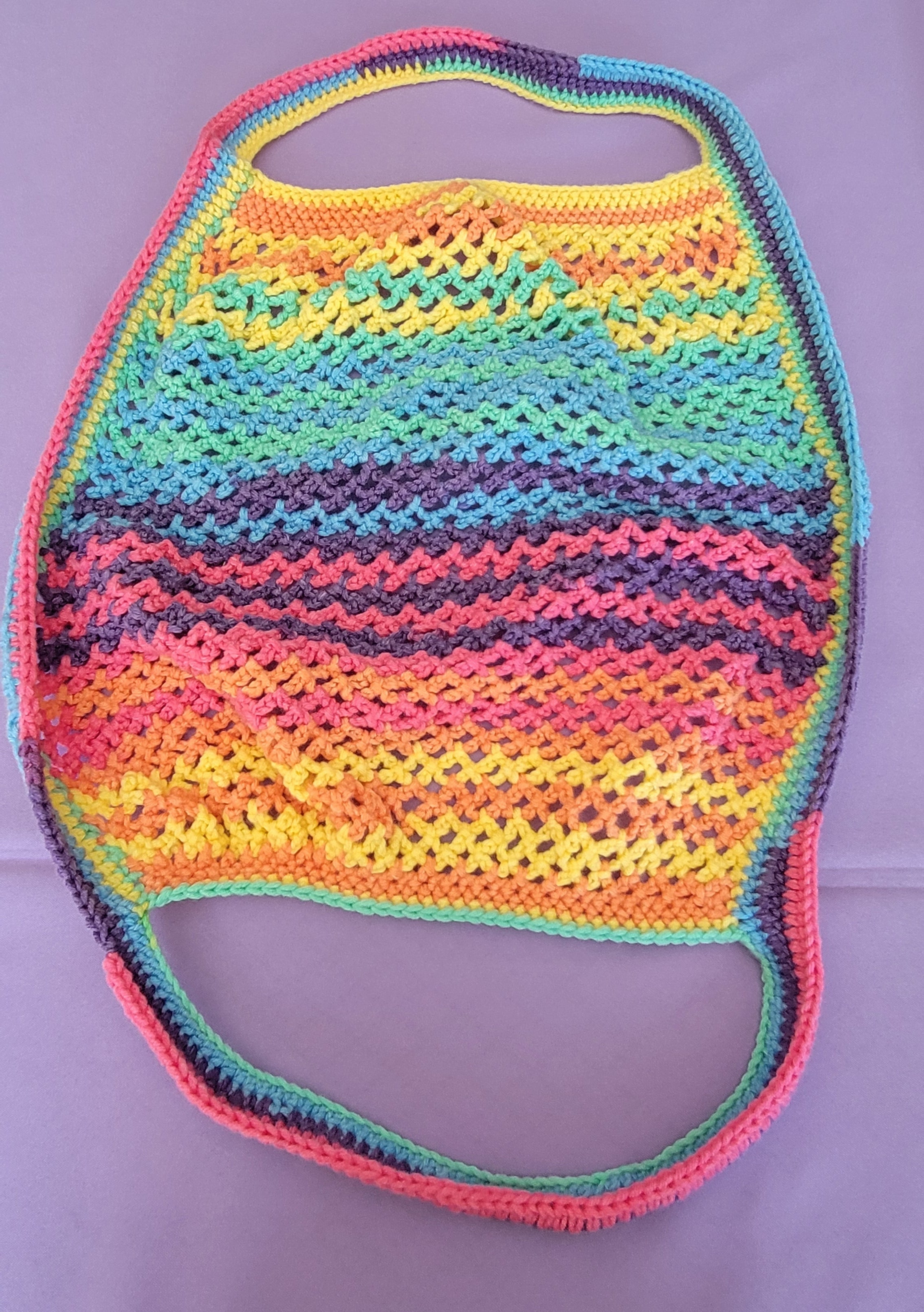 A handmade crochet shoulder bag with a vibrant rainbow stripe pattern, laid flat on a light purple background.