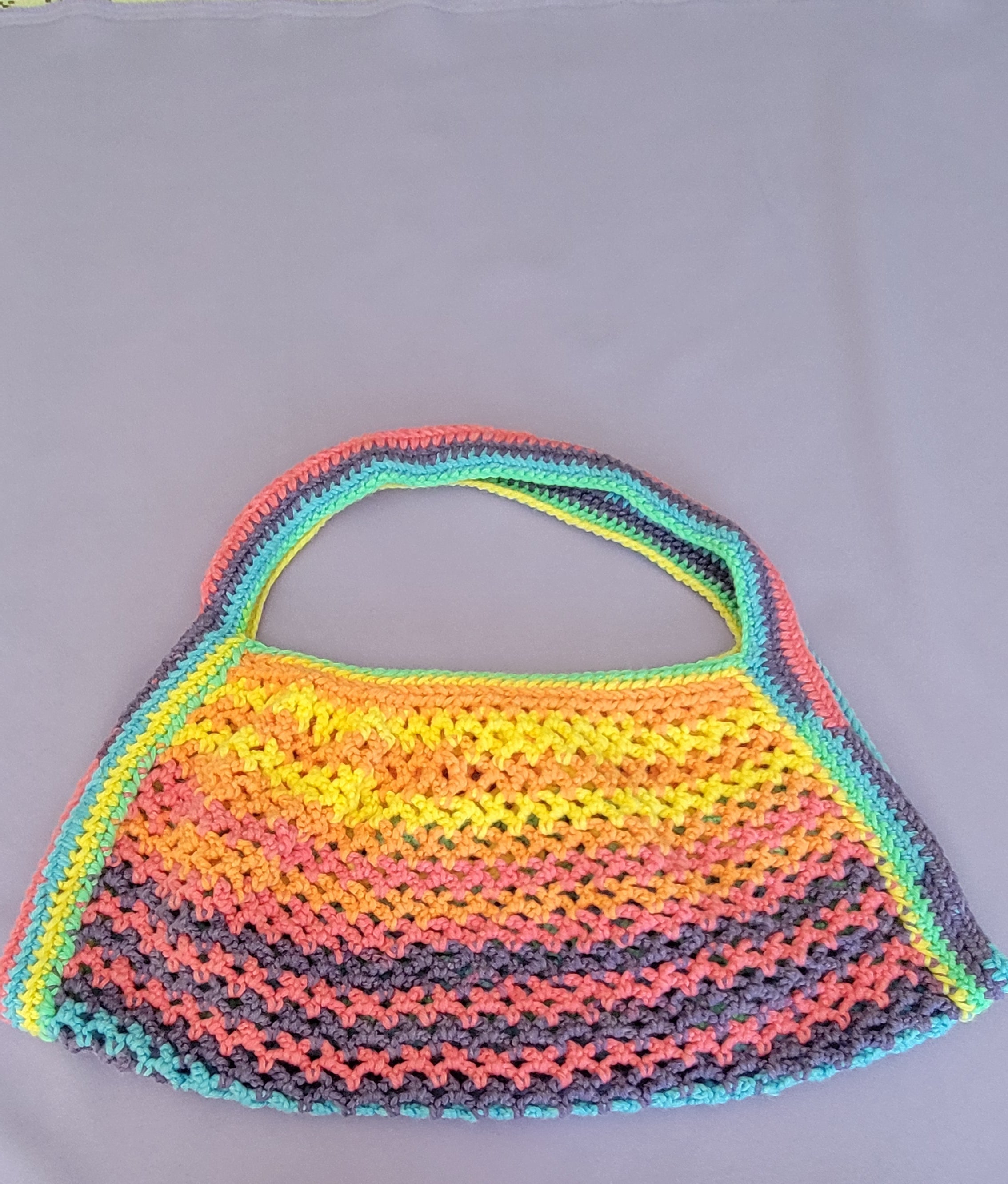 A handmade crochet shoulder bag with a vibrant rainbow gradient pattern of yellow, orange, pink, purple, and blue, featuring a single top handle, laid flat on a gray background.