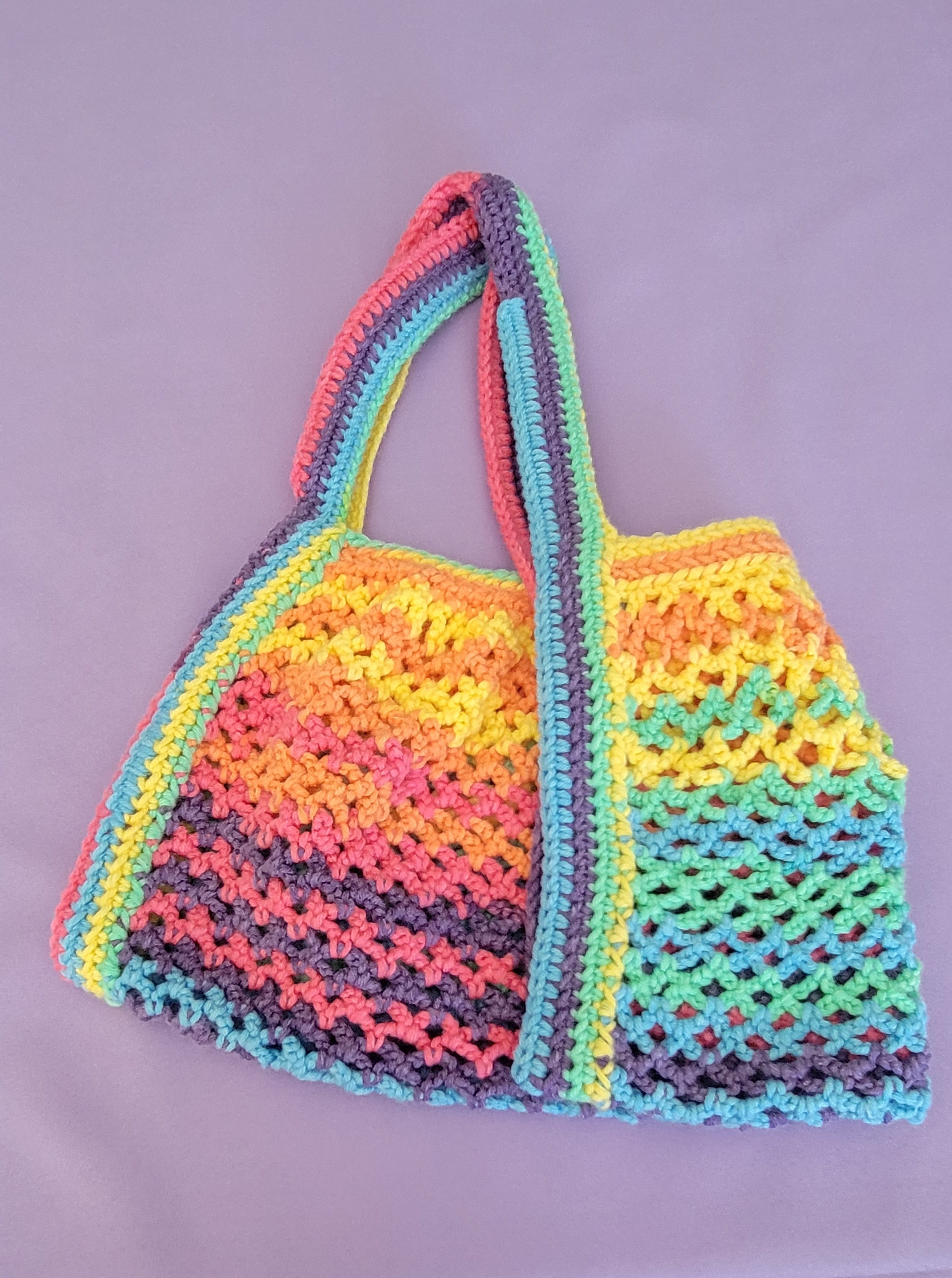 A handmade crochet shoulder bag with a vibrant rainbow pattern of pink, orange, yellow, green, blue, and purple, laid flat on a light purple background.