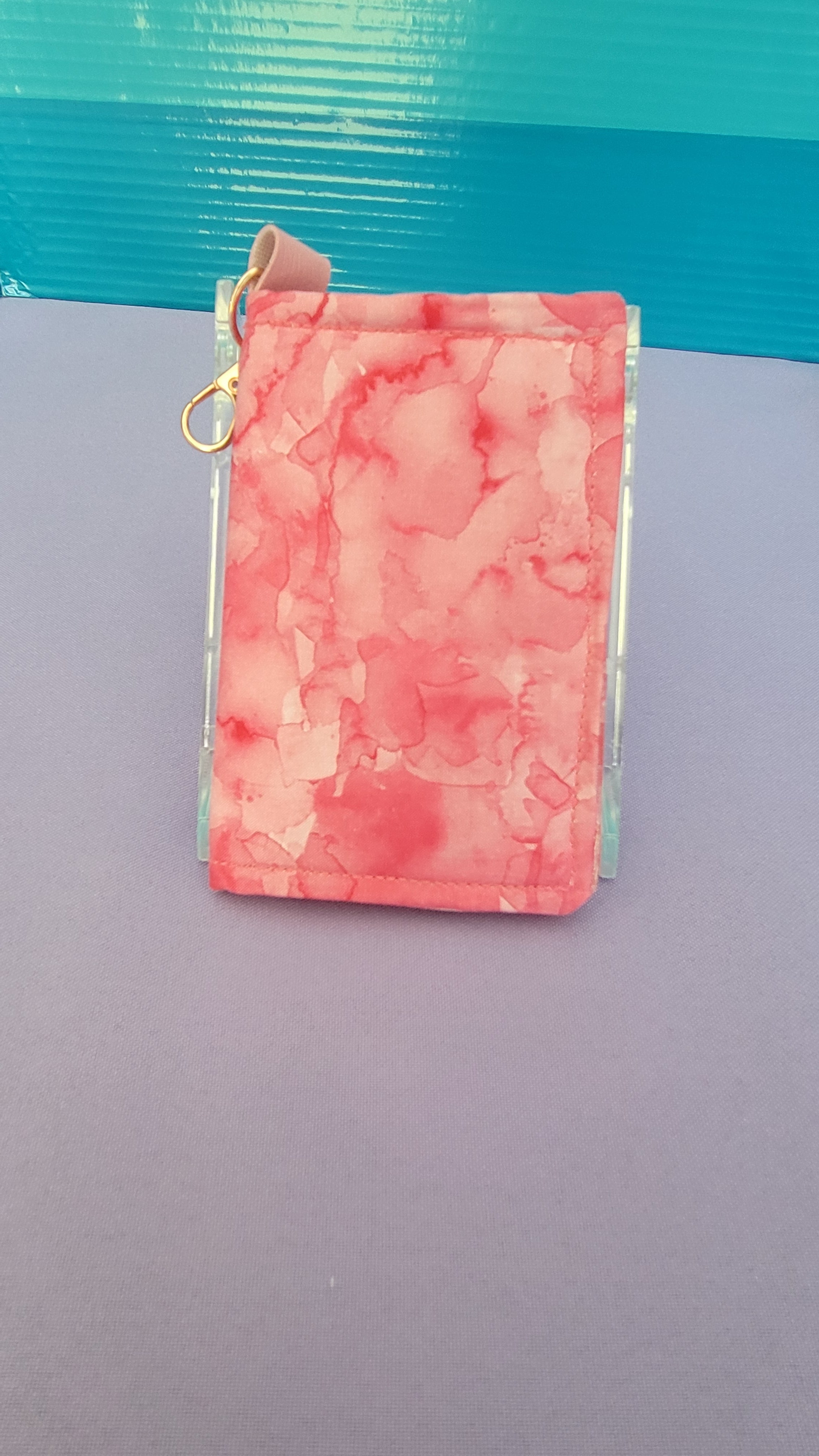 A small wallet with a pink and red marbled pattern, featuring a small tan leather loop and a gold metal ring, shown on a split purple and teal background.