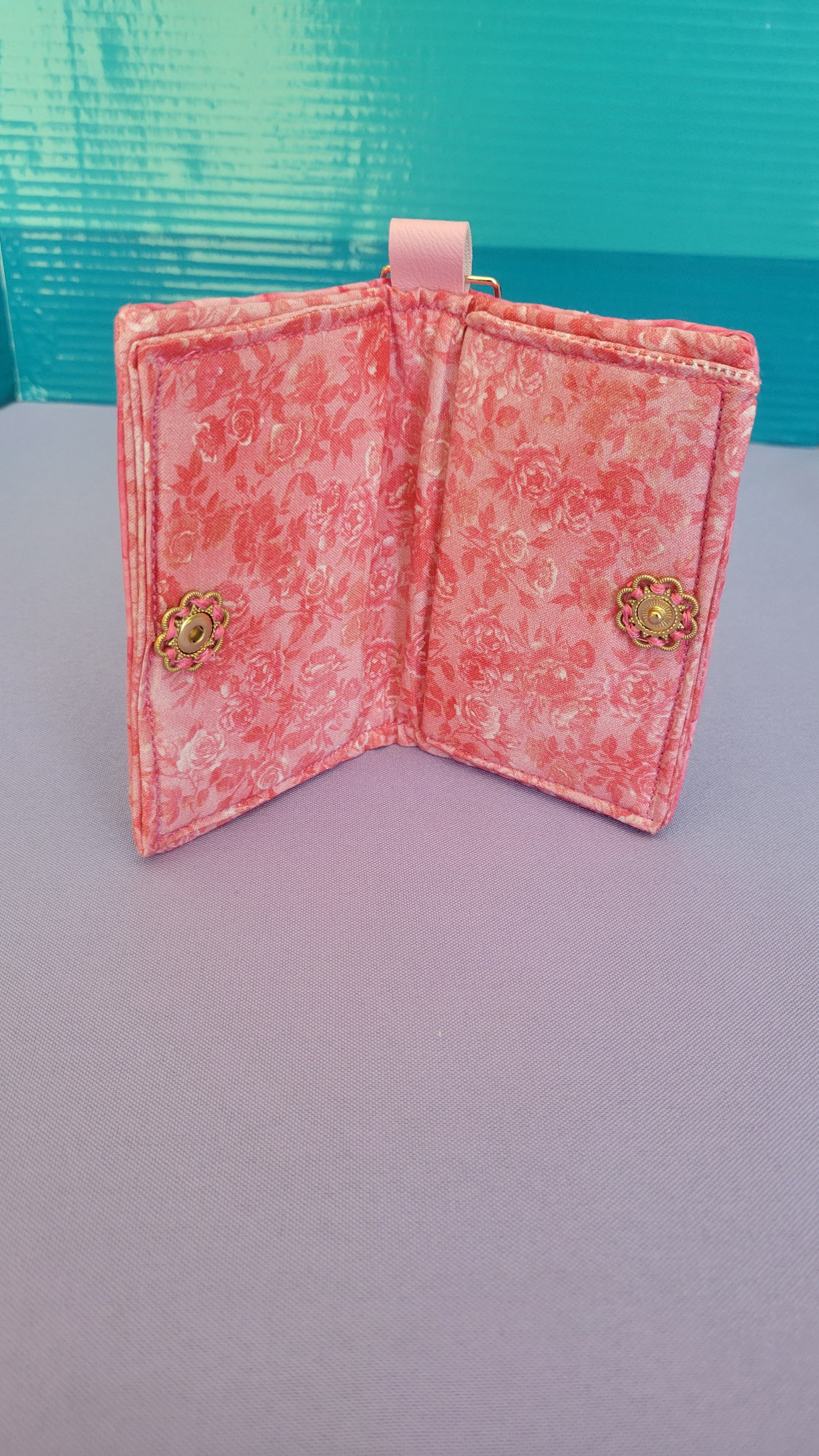 A pink floral fabric wallet with gold-tone metal snap closures, shown open against a split background of light purple and teal.