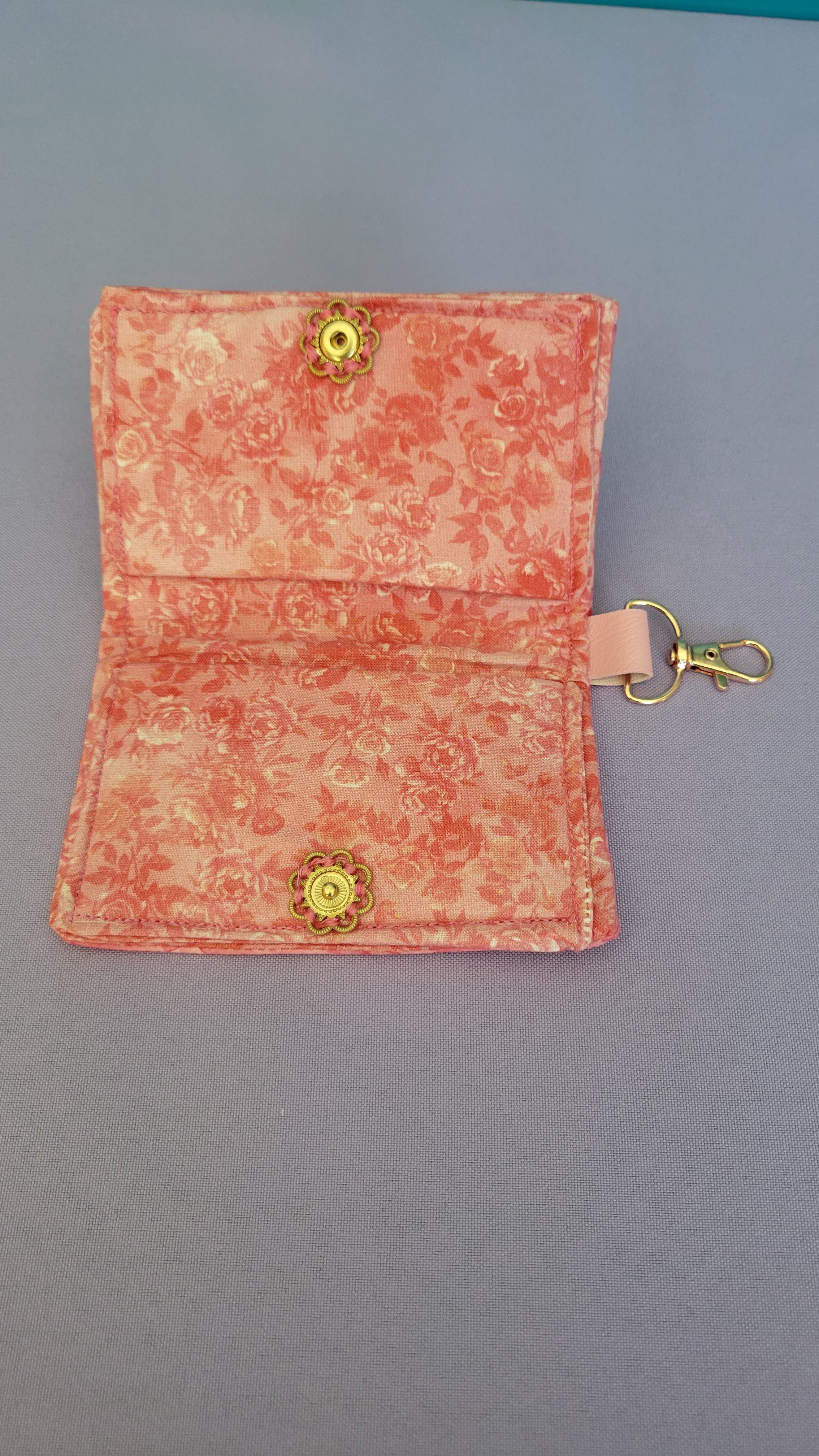 A small fabric wallet or card holder in a coral pink and beige floral pattern, featuring two gold-tone snap buttons and a gold key ring clasp at the top, shown on a plain gray background.