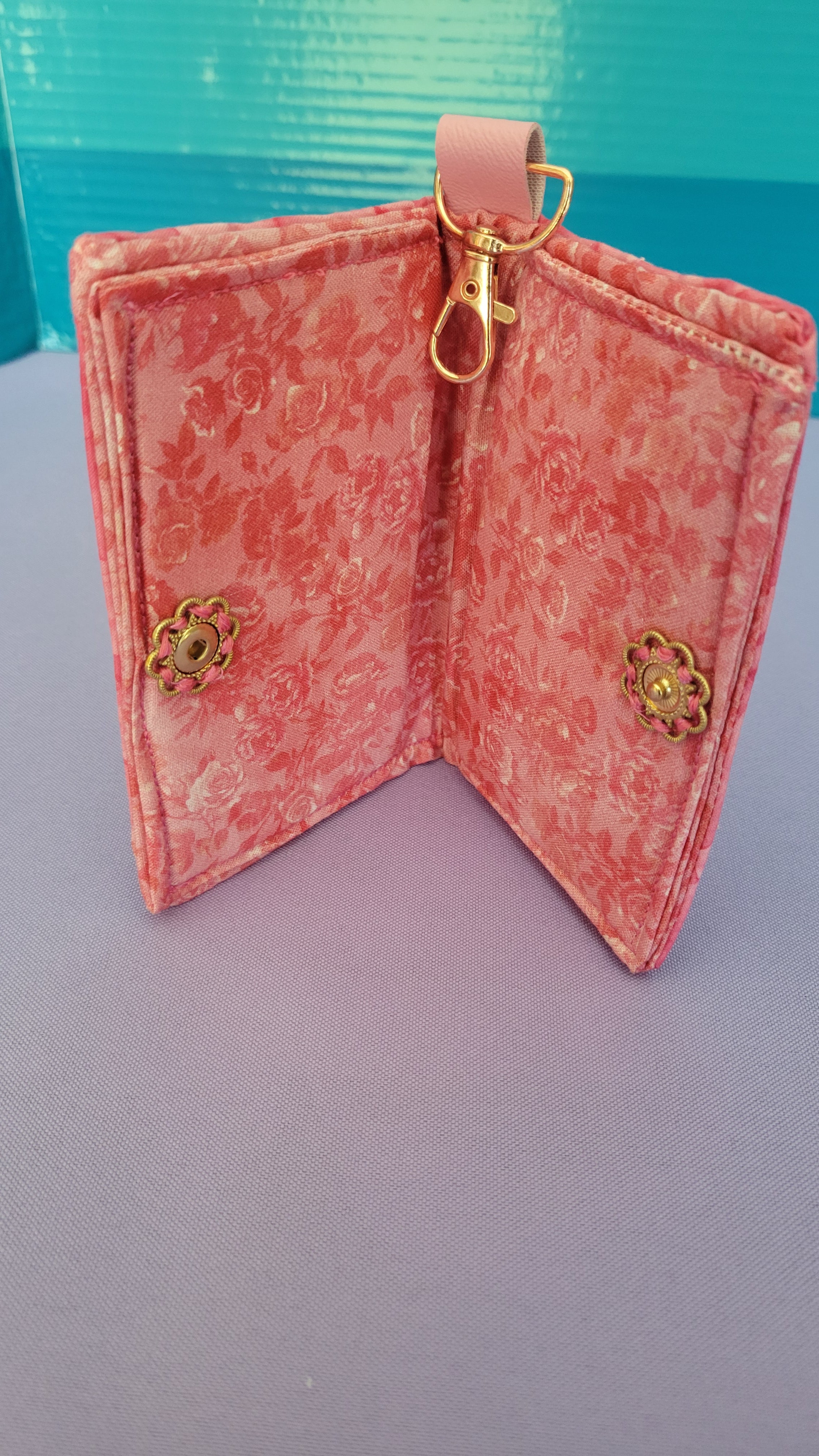 A pink floral patterned wallet with gold-tone vitange flower push buttom, shown open on a split purple and blue background.