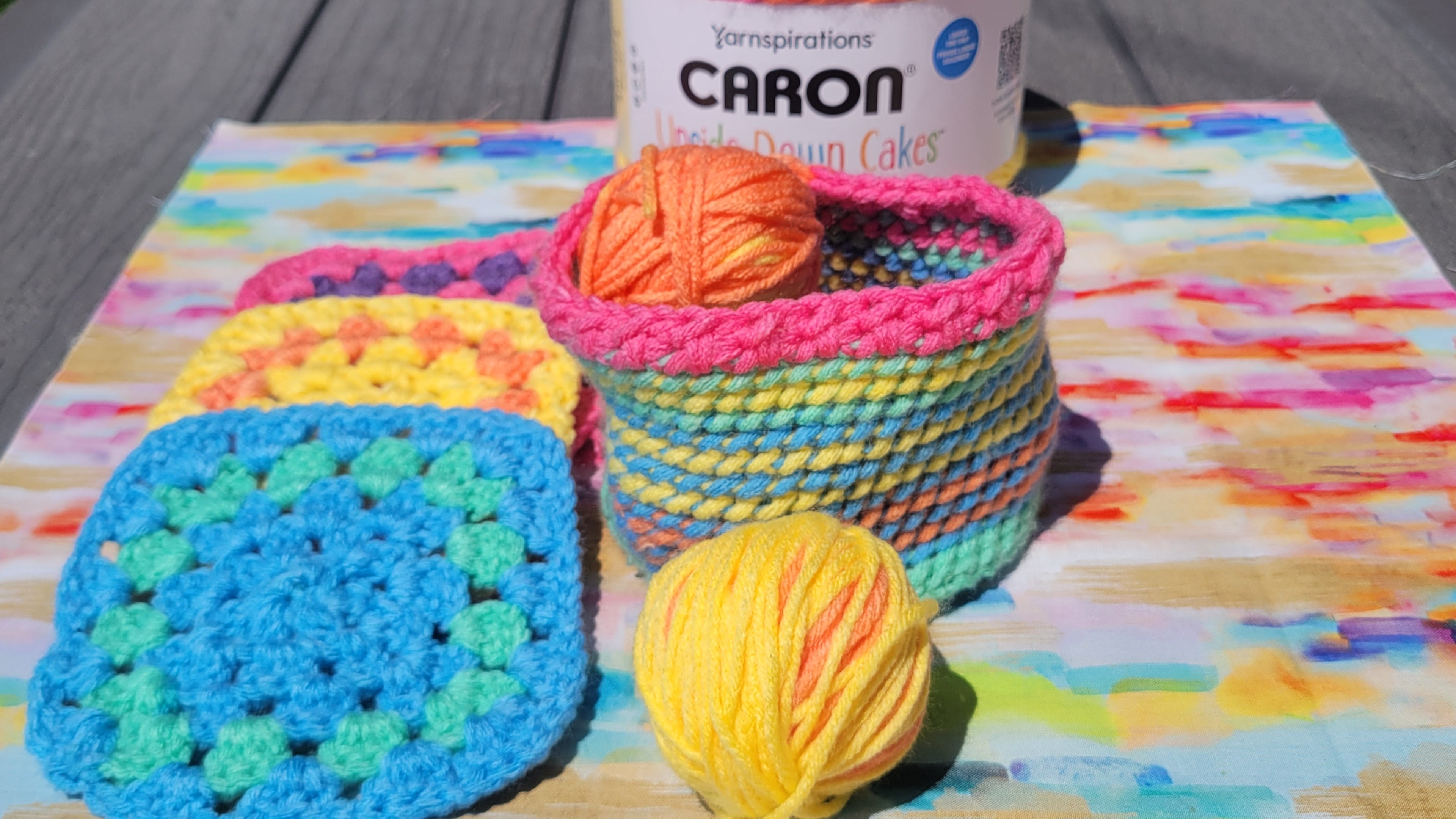 A colorful crocheted yarn storage basket with striped pink, yellow, blue, and green bands, holding a ball of orange yarn. A matching ball of yellow and pink yarn sits in front, with folded crocheted square samples and a canister labeled Yarnspirations CARON on a painted paper backdrop.
