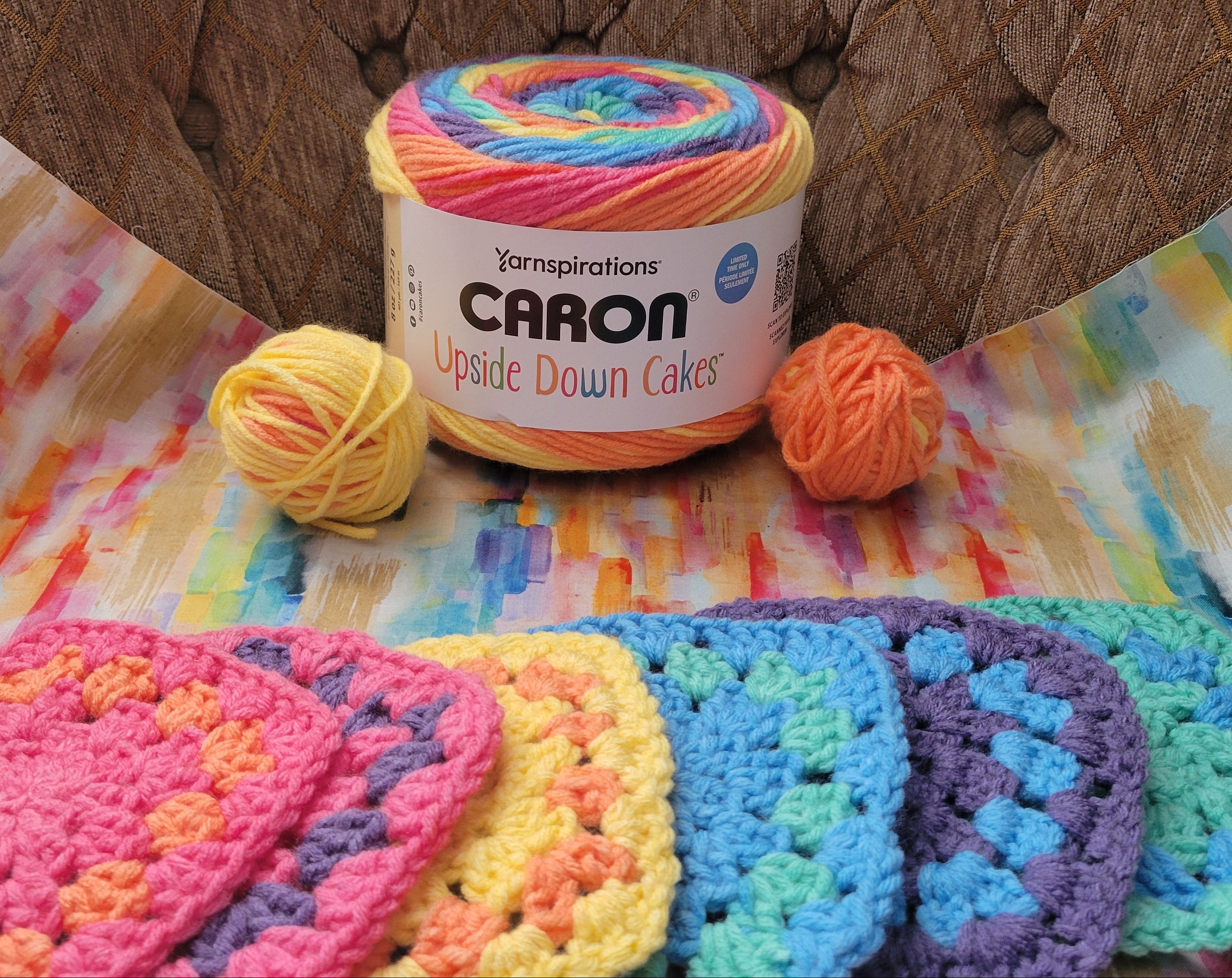 A yarn skein labeled Yarnspirations Karon Upside Down Cakes sits on top of a colorful printed sheet, with two small balls of yarn beside it and several crocheted swatches in pink, yellow, blue, purple, and green arranged in front. The setup is placed on a brown tufted chair.