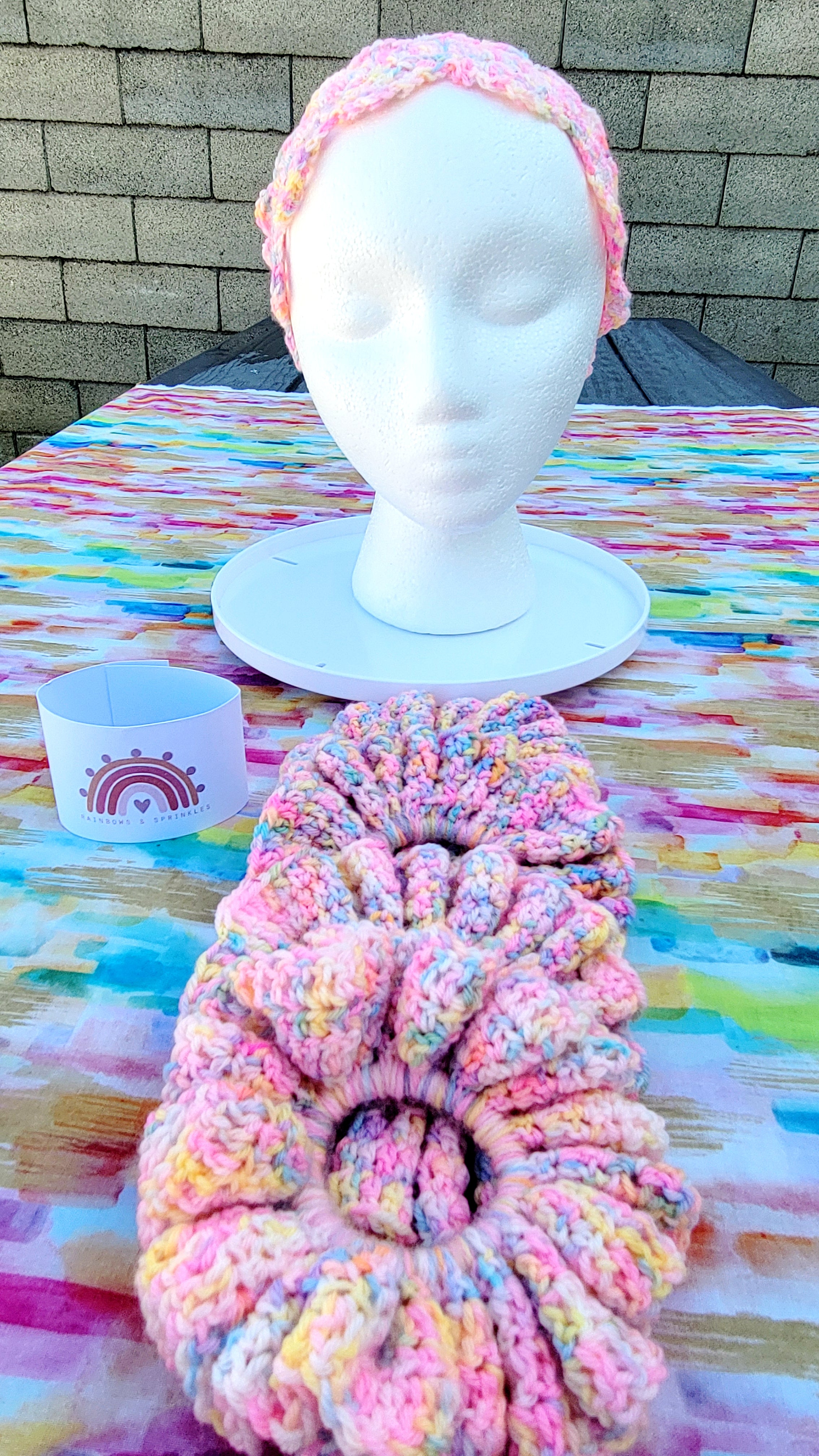 Four crochet scrunchies in a variegated pastel pink, yellow, and blue yarn is displayed in the foreground on a colorful painted table. In the background, a matching crochet headband is shown on a white mannequin head. Next to the item is the promotion of Rainbow and Sprinkles.