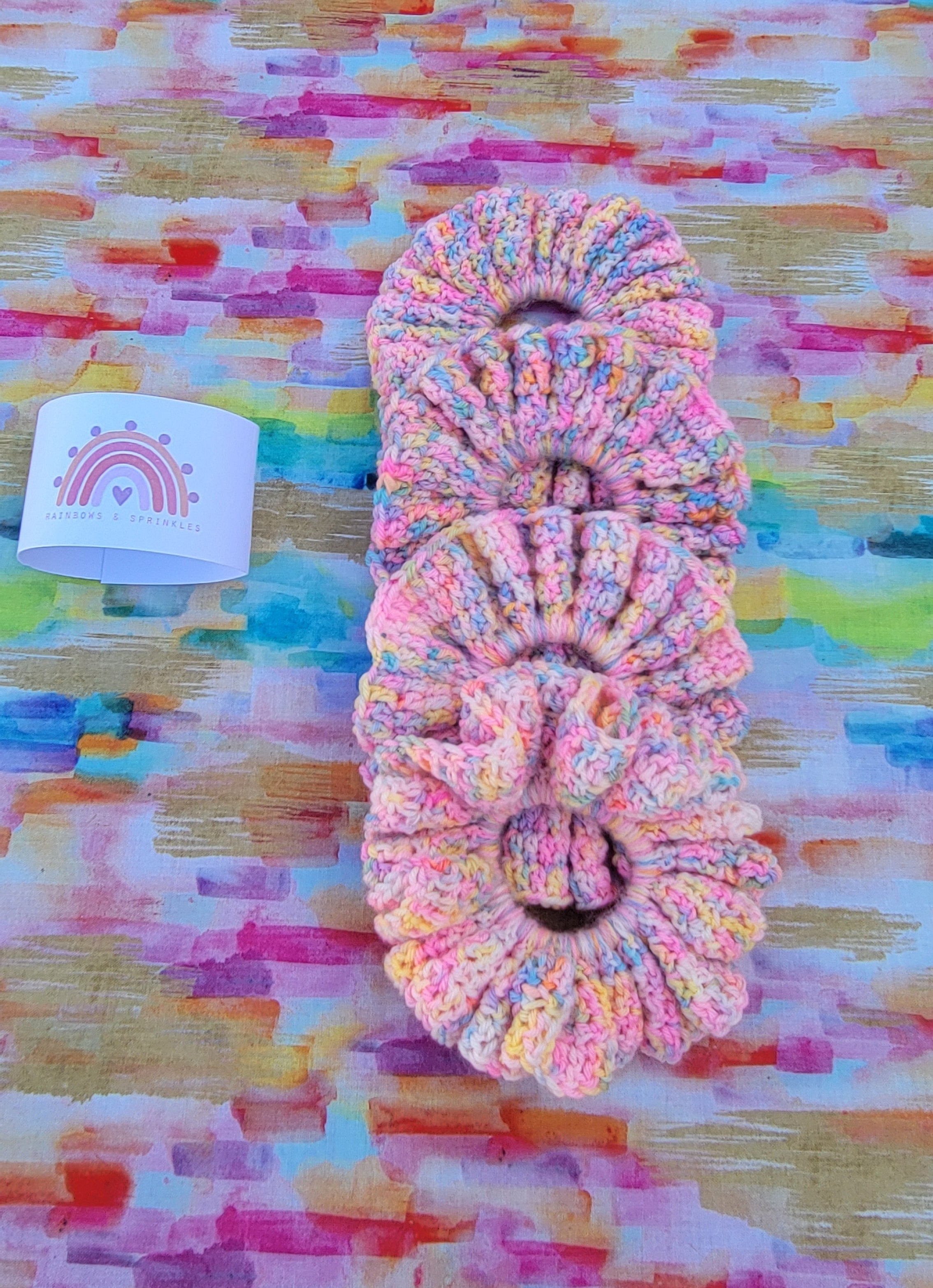 Four crochet pastel hair scrunchies stacked vertically on a colorful abstract painted background, with a small white paper tag showing a rainbow logo to the left.