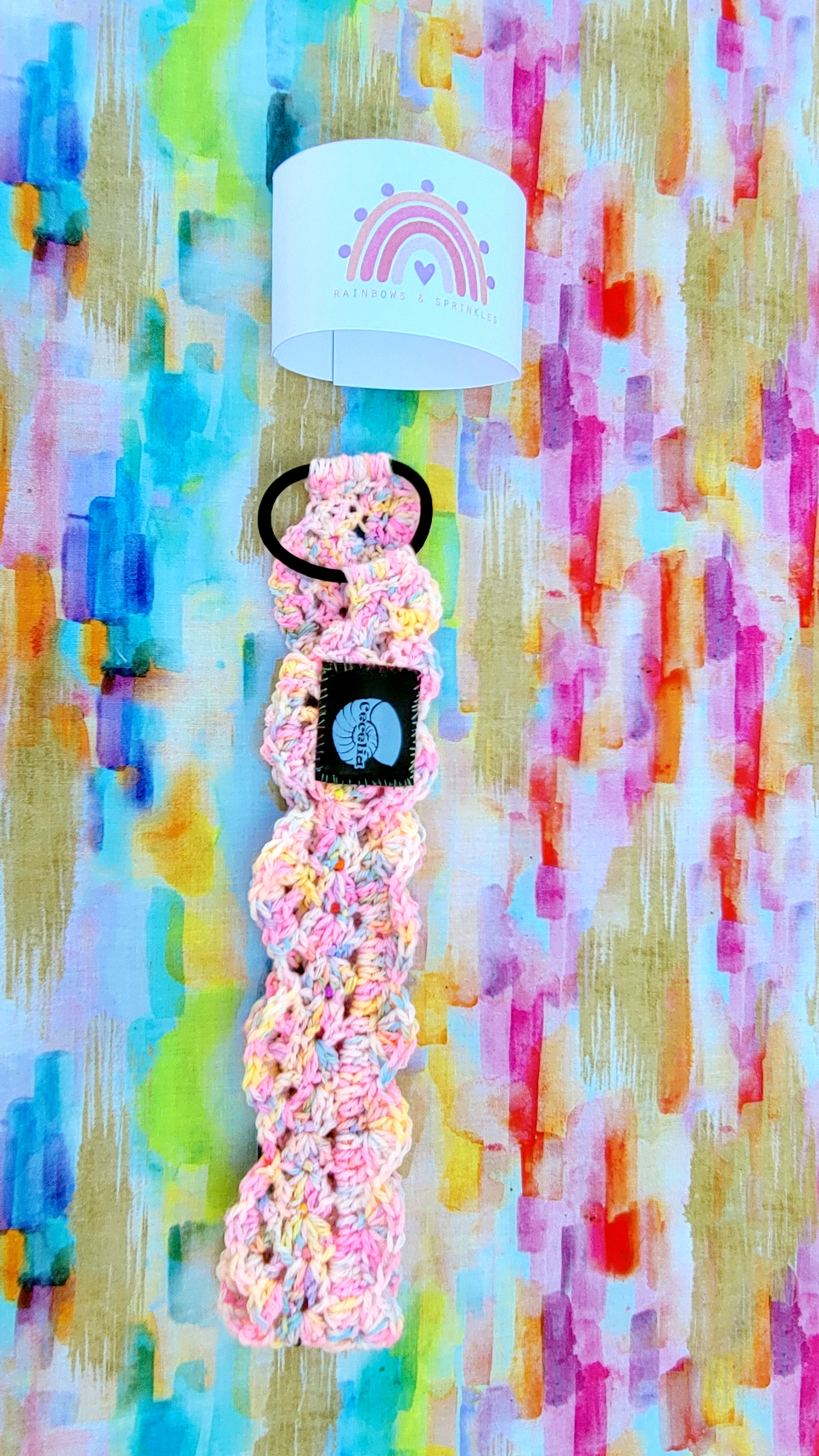 A pastel pink and yellow crocheted  hairband with a black rubber band and a black square patch featuring the brands name, displayed against a colorful abstract painted background with a white cylindrical display stand. On top of the item is the promotion of Rainbow and Sprinkles.