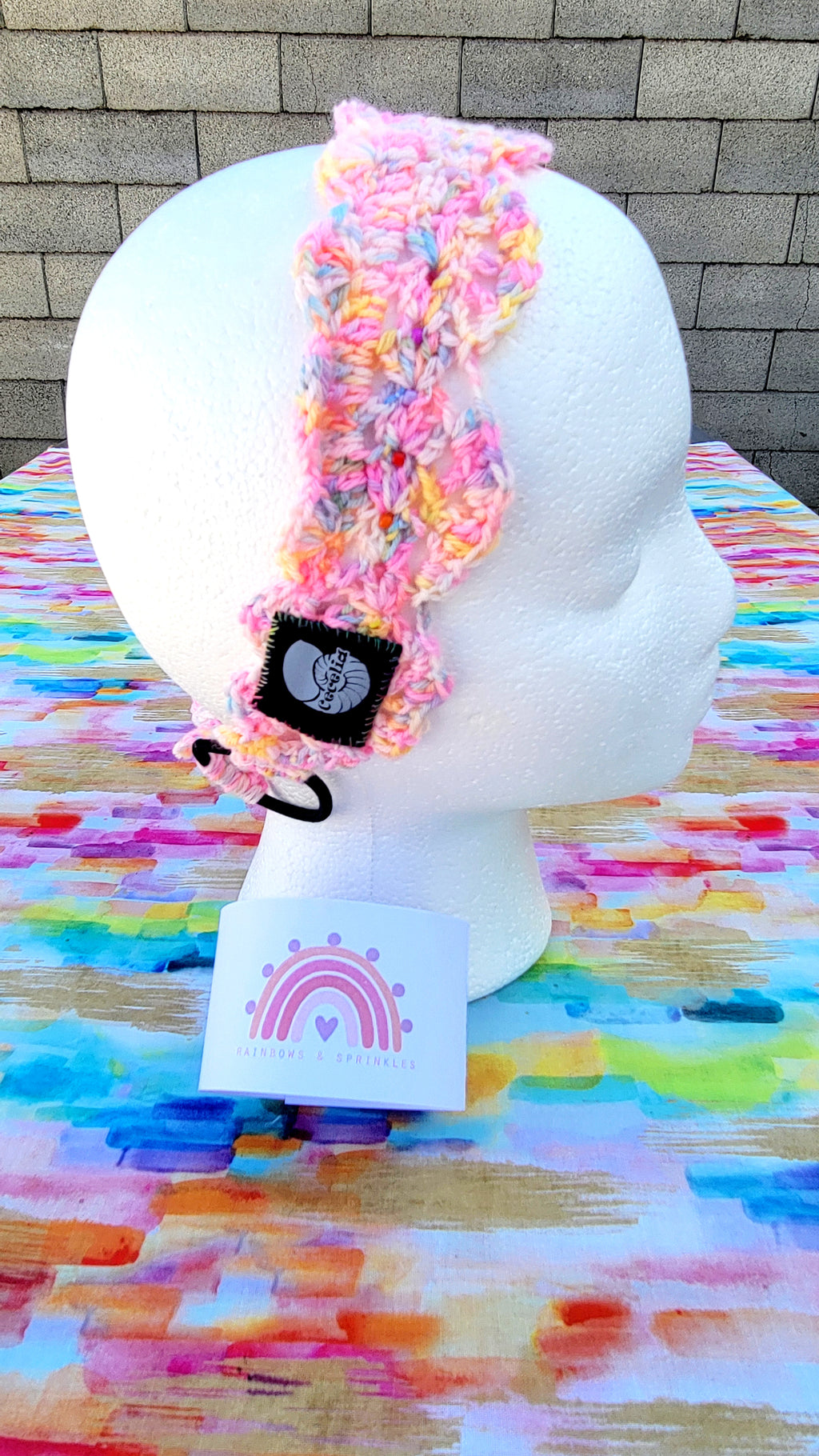 A multicolor pastel crochet headband with glass beads handsewn, a black fabric patch featuring a white sea shell and the text 'Cecelia Headband', displayed on a white styrofoam mannequin head. The headband is resting on a colorful painted table surface with a gray brick wall in the background. On the bottom of this item is the promotion of Rainbow and Sprinkles.