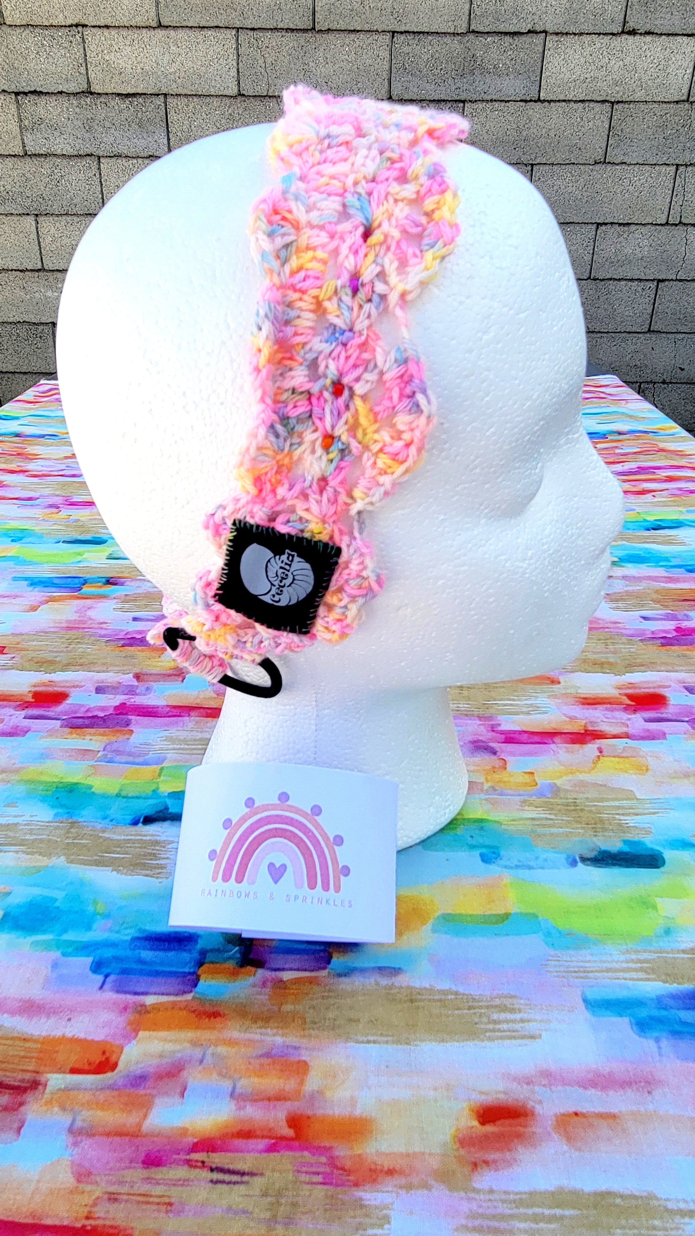 A multicolor pastel crochet headband with glass beads handsewn, a black fabric patch featuring a white sea shell and the text 'Cecelia Headband', displayed on a white styrofoam mannequin head. The headband is resting on a colorful painted table surface with a gray brick wall in the background. On the bottom of this item is the promotion of Rainbow and Sprinkles.