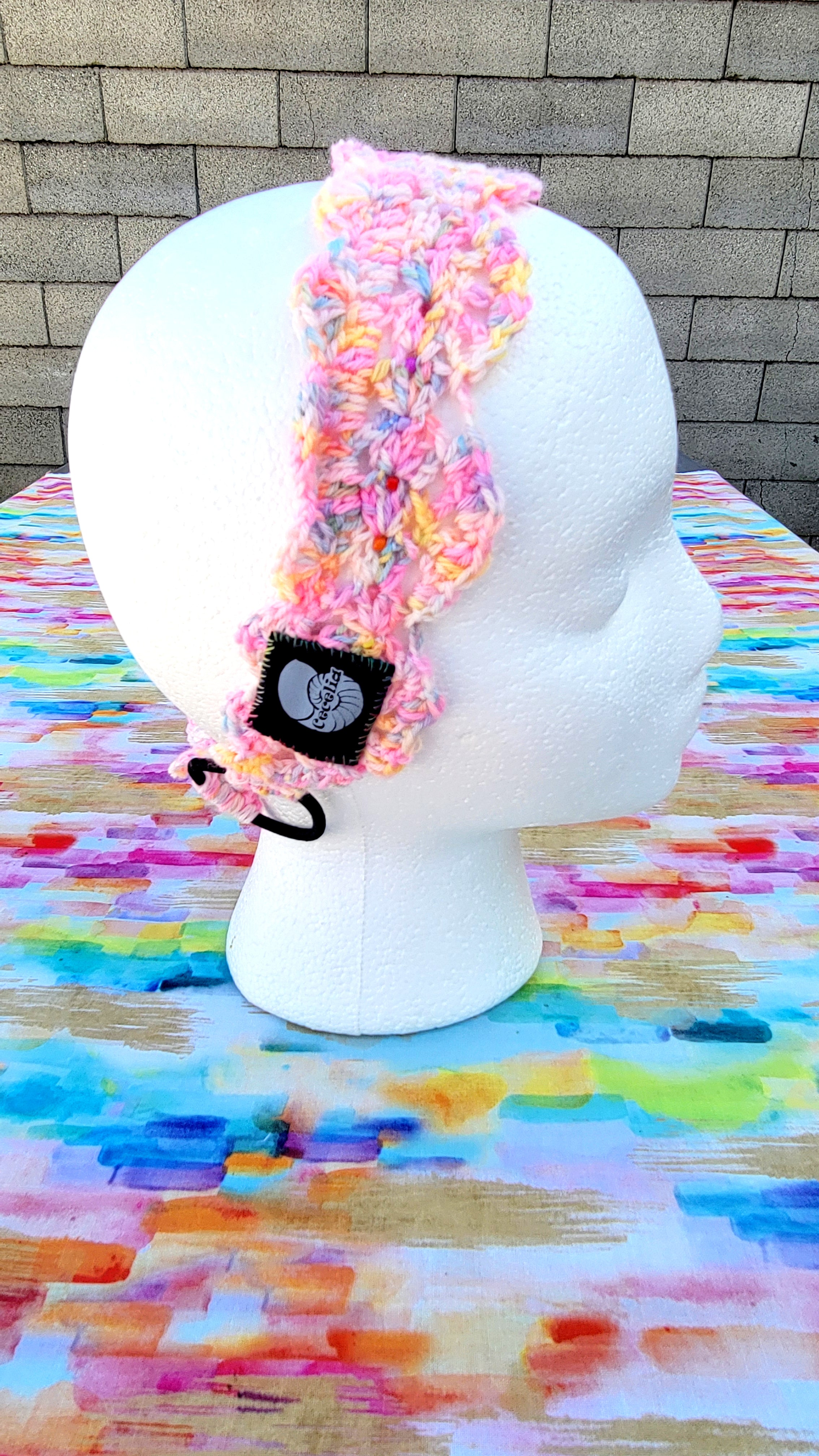 A handmade crocheted hair in a variegated pink, yellow, and orange yarn pattern, displayed on a white styrofoam mannequin head. The headband features handsewn glass beads, a black fabric patch with a gray circular graphic with a white shell and the brand name 'Cecelia'