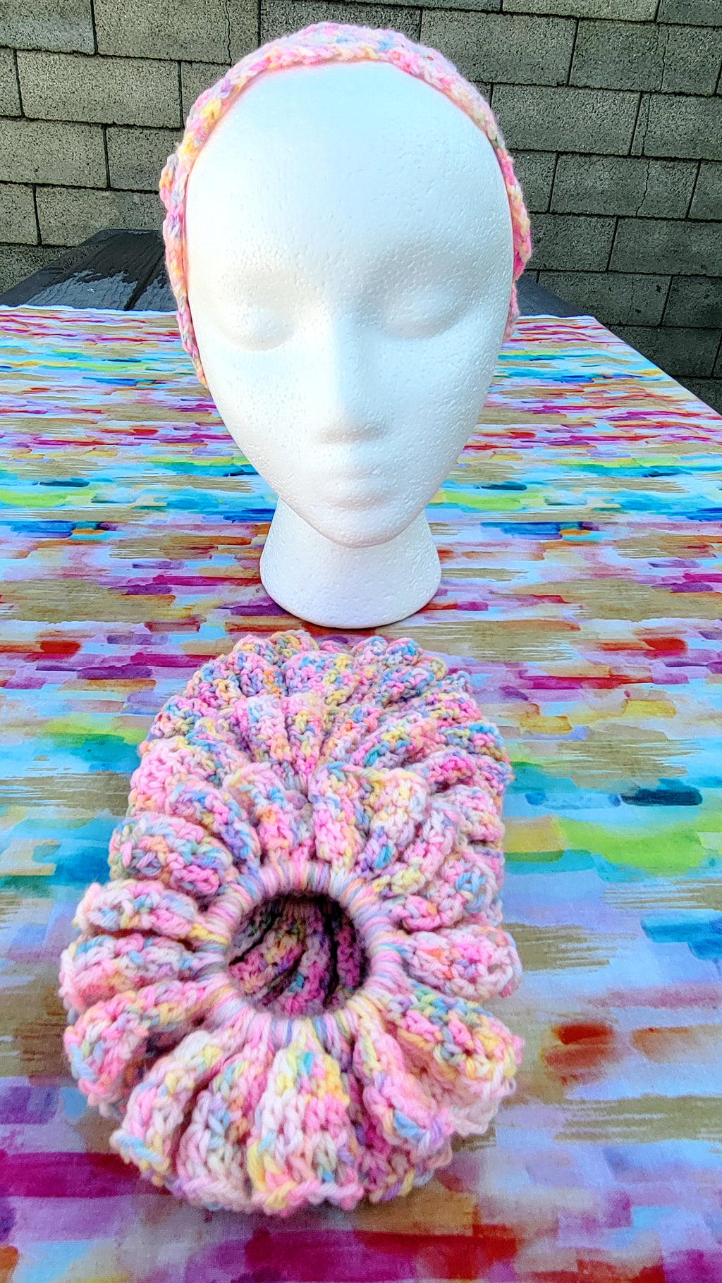 four crochet scrunchies in variegated pink, yellow, and blue yarn sits on a colorful painted table surface, with a white mannequin head wearing a matching pink hairband behind it.