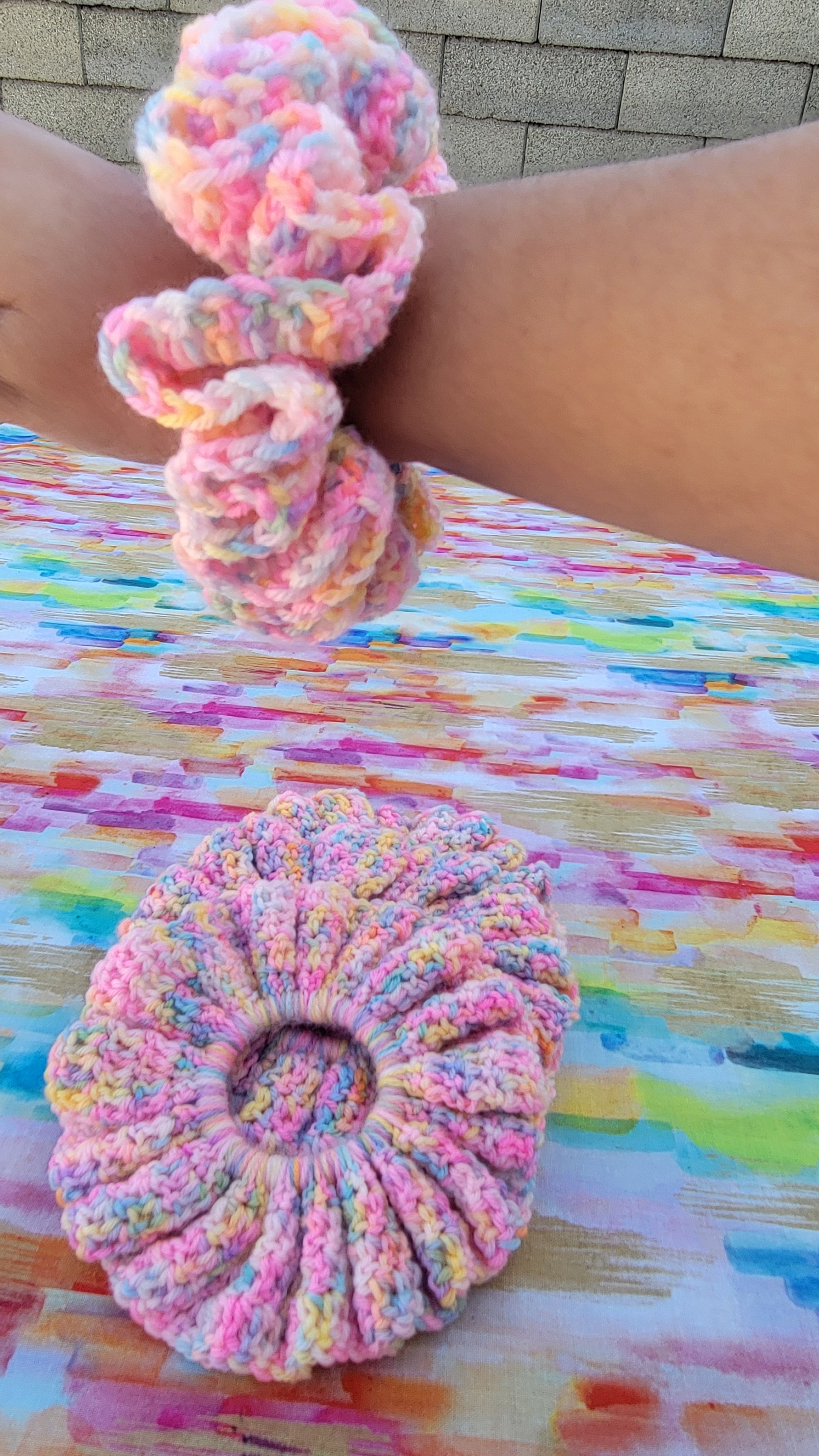 Four chunky hand-crocheted scrunchies in a pastel variegated pink, yellow, and blue yarn, shown with one on a person's wrist and one laid flat on a colorful painted wooden table.