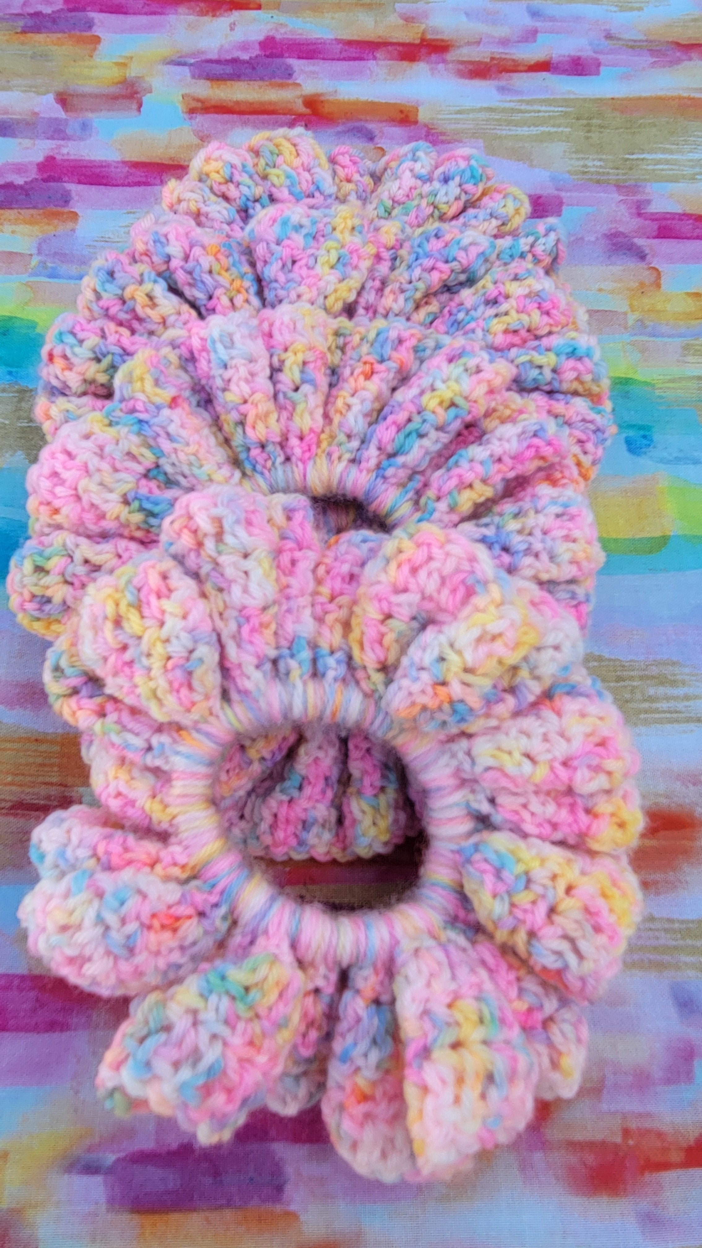 Four large, chunky hand-crocheted hair scrunchies in a pastel variegated yarn blend of pink, yellow, blue, and white, displayed on a colorful painted wood background.