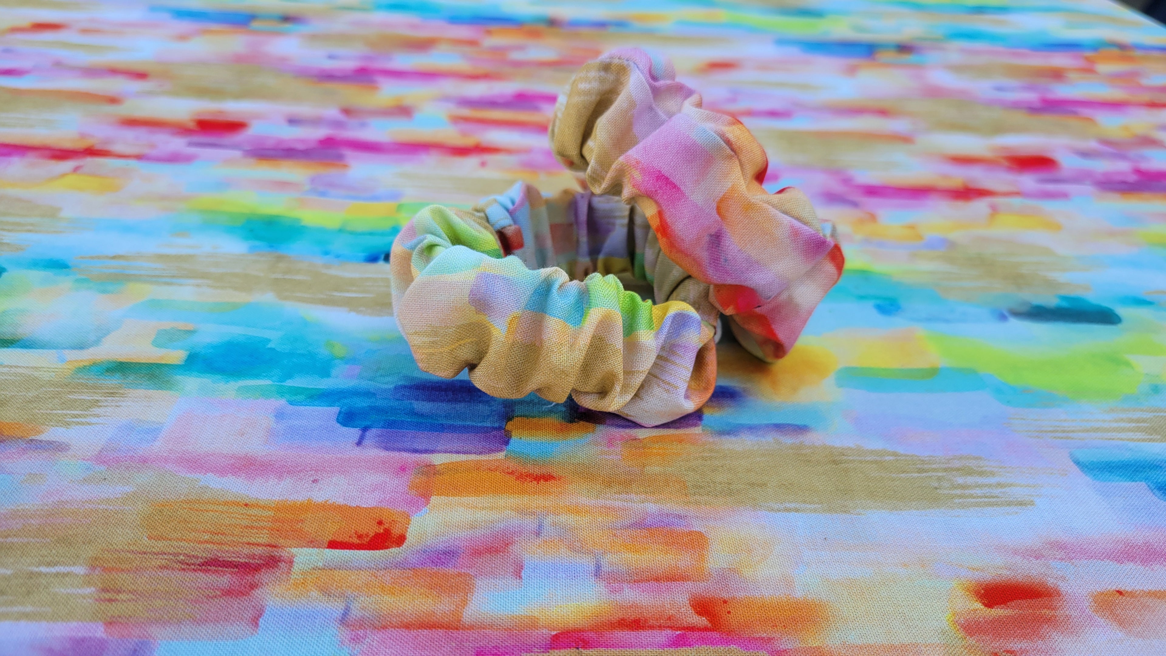 Two fabric scrunchies in pastel rainbow colors resting on a colorful painted surface.