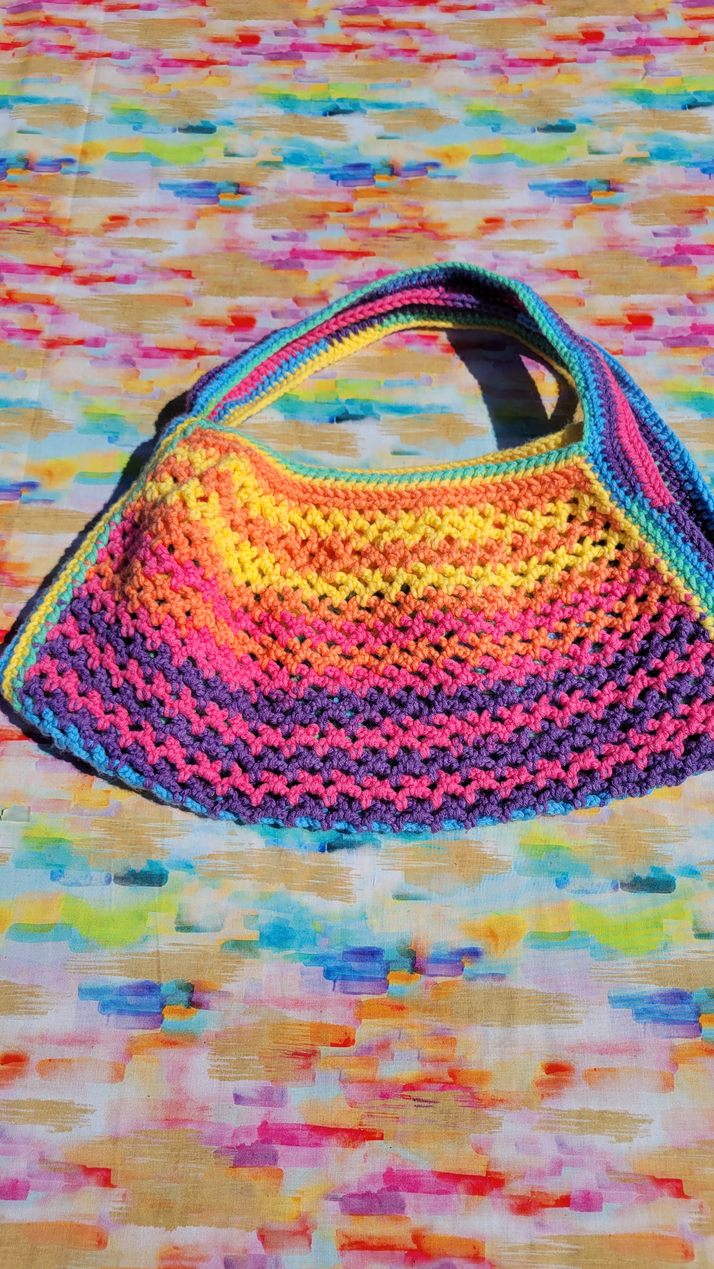 A handmade crocheted shoulder bag with a vibrant rainbow gradient of pink, yellow, orange, green, and blue, laid flat on a colorful abstract patterned surface.