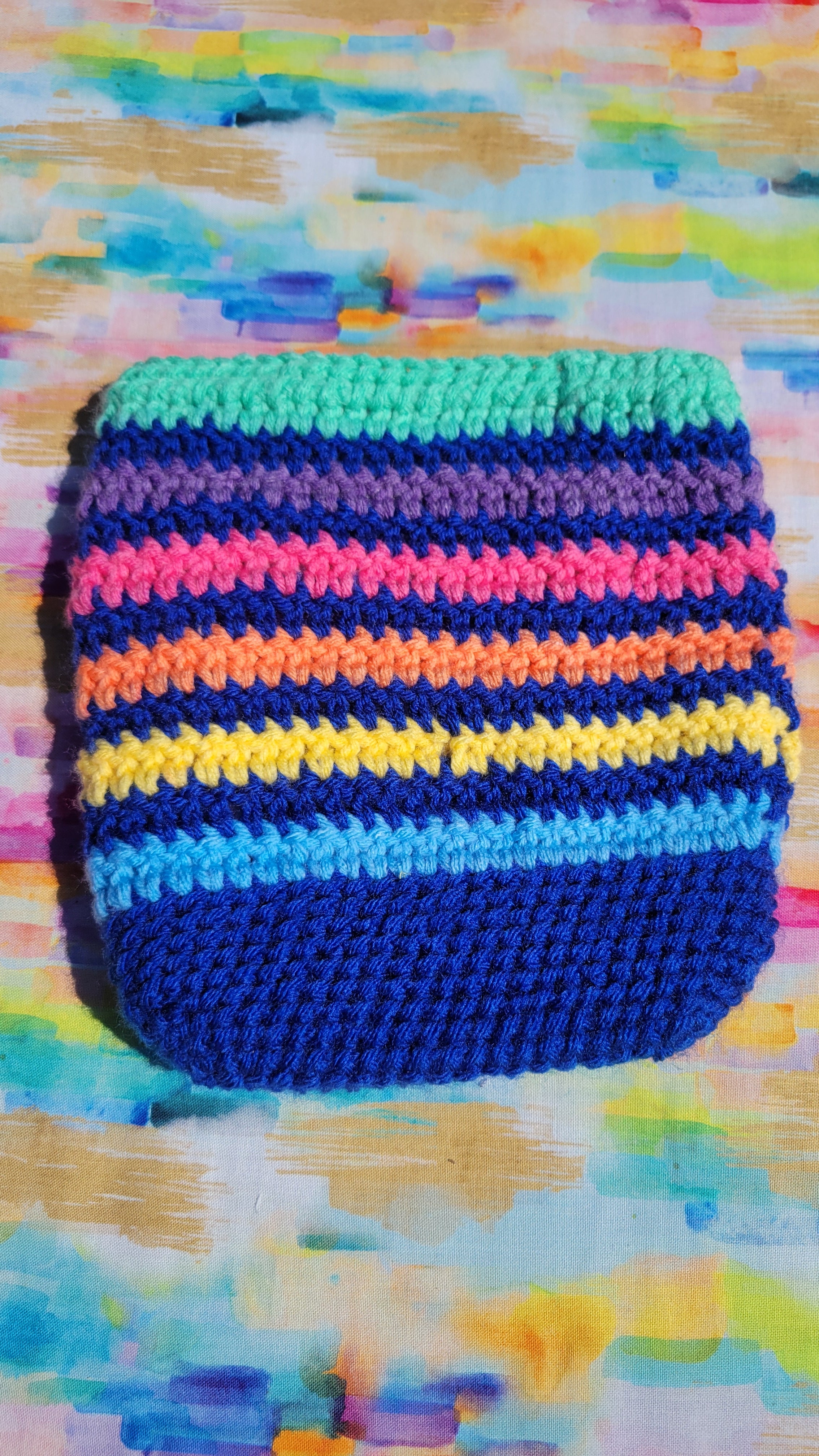 A handmade royal blue crocheted makeup bag with horizontal stripes in green, purple, pink, orange, yellow, and blue, placed on a colorful painted background.