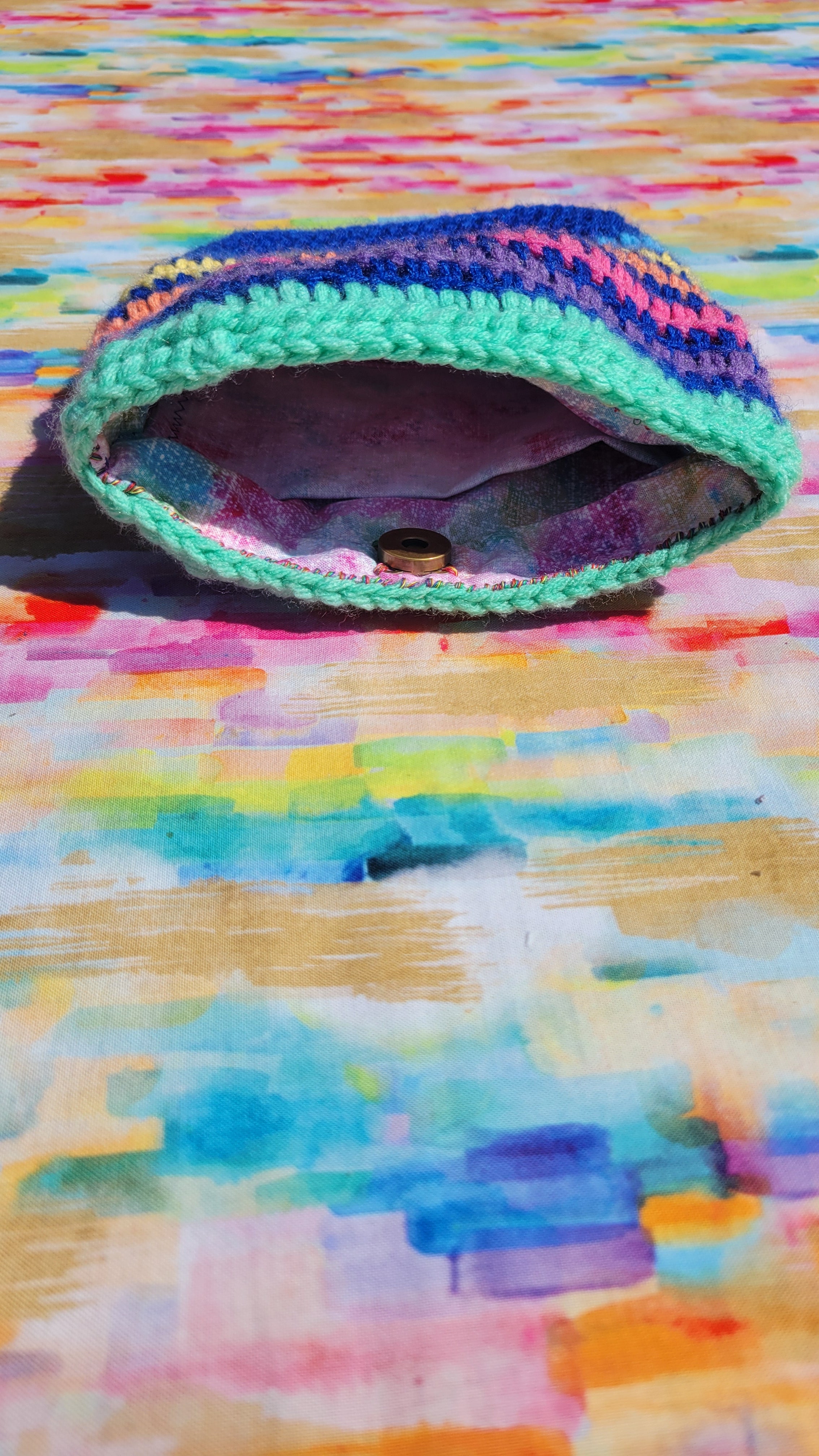 A handmade crocheted royal blue makeup bag with a colorful variegated pattern of purple, blue, green, and pink, featuring a green trim and a snap button in a broze color closure, resting on a vibrant, multicolored painted background.