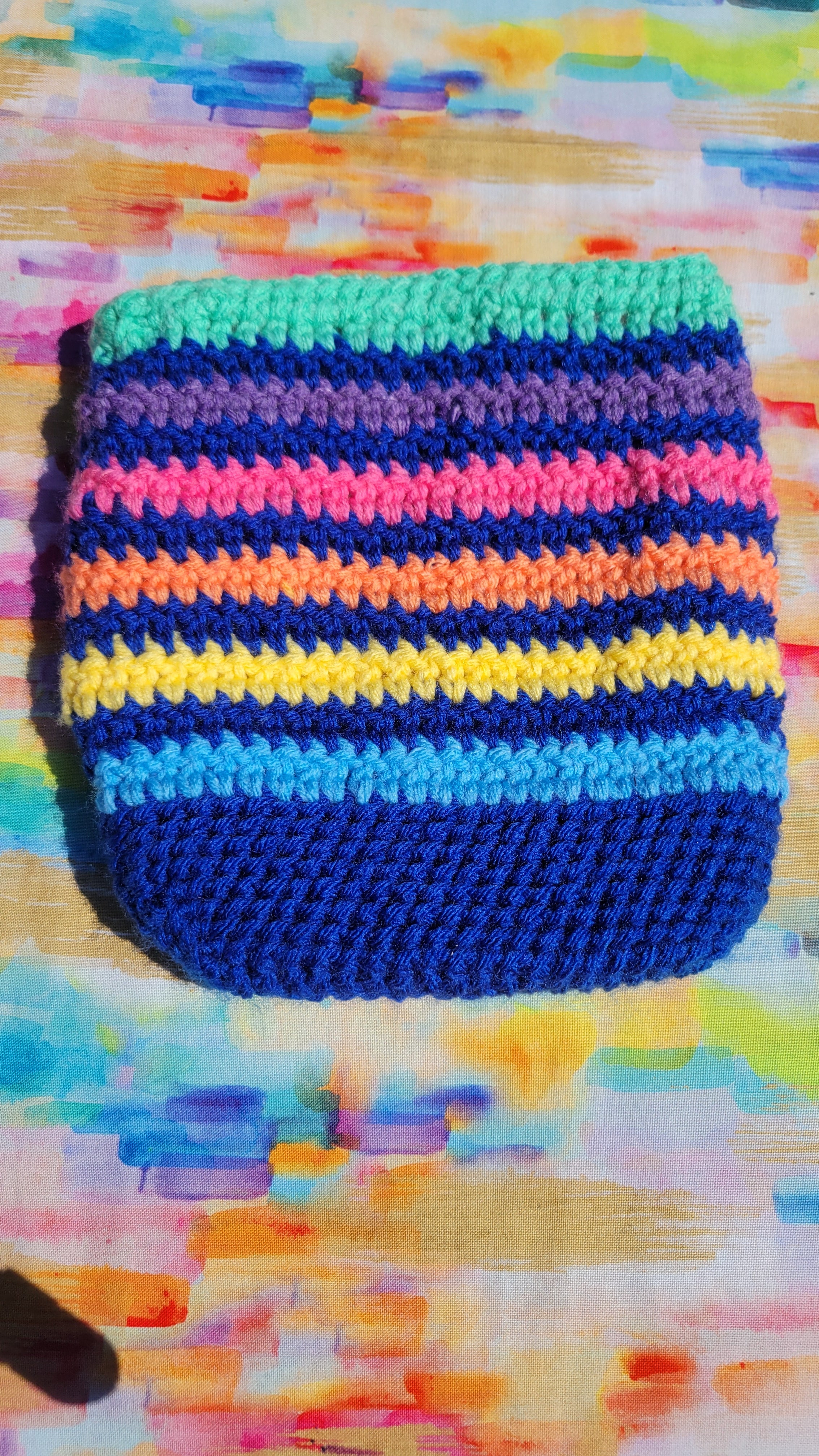 A handmade crocheted royal blue makeup purse with horizontal stripes in green, purple, pink, orange, yellow, and light blue, resting on a colorful painted canvas background.