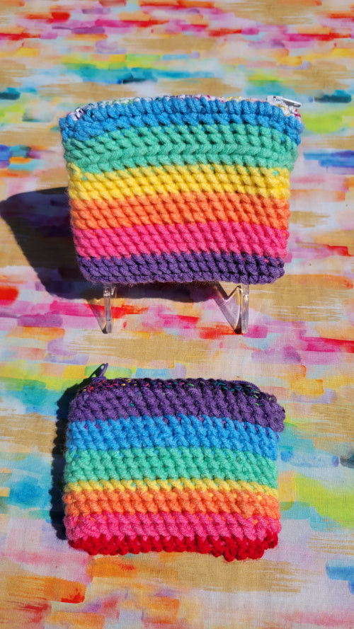 Two small hand-crocheted zippered pouches for cards with rainbow-colored stripes, displayed on a colorful painted background.