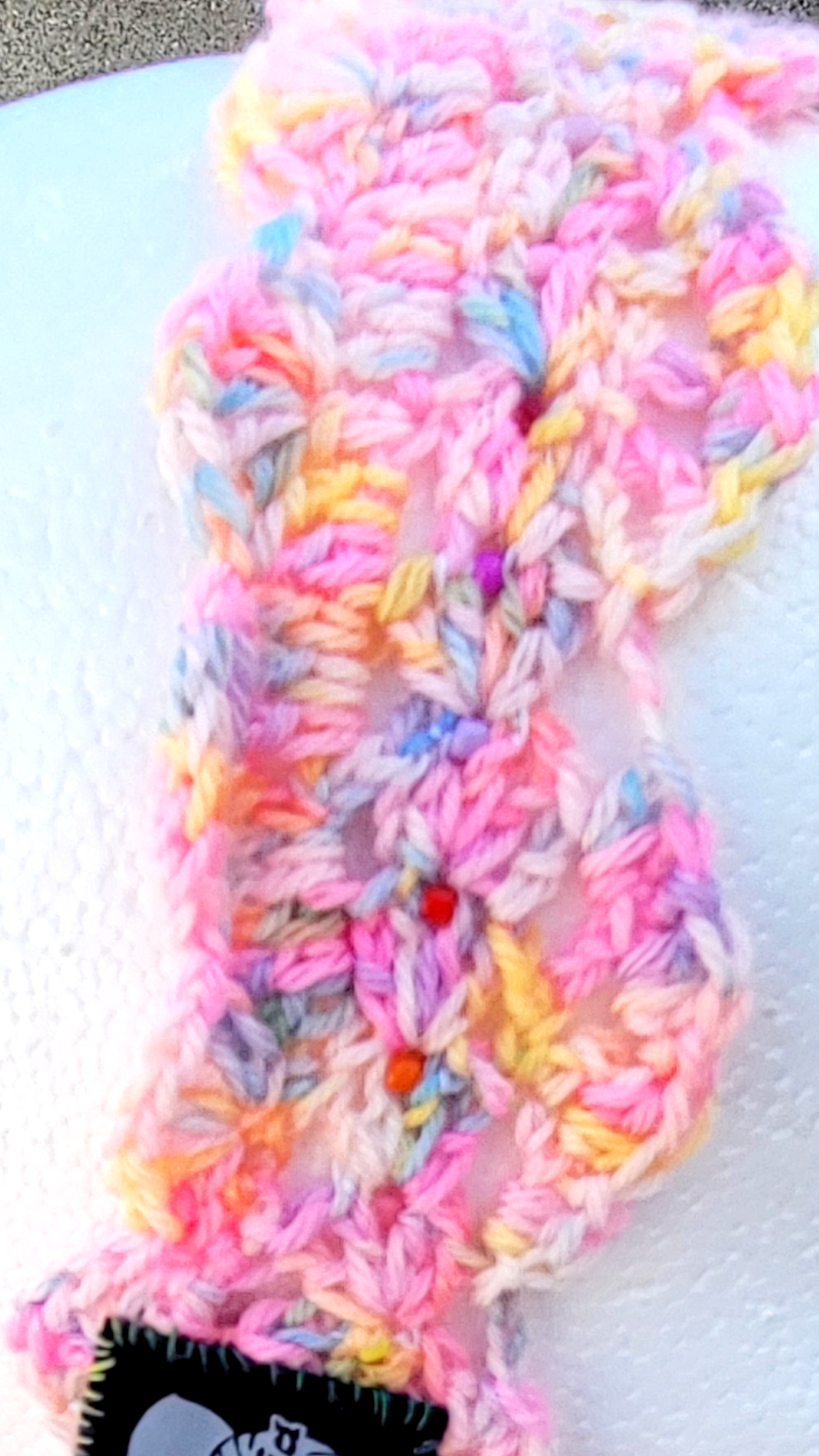 A close-up of a handmade crochet accessory, hairband made from glass beads, variegated yarn in pastel pink, yellow, blue, and white. A black plastic pin is threaded through the center to secure the stitches, and a black fabric label is visible at the bottom.