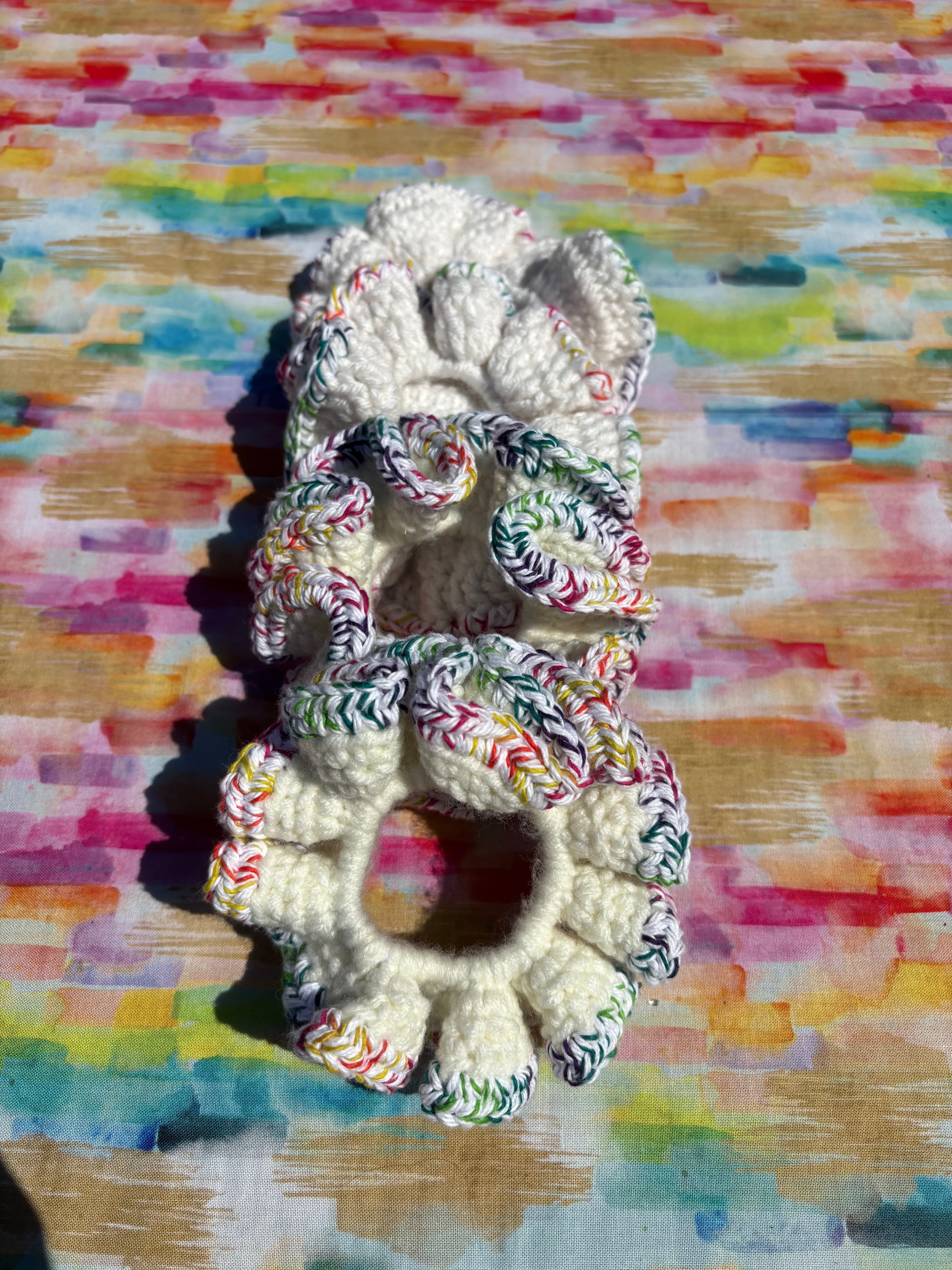 Four handmade crocheted scrunchies vertically, featuring a cream-colored center in white or off-white with multicolored striped fabric accents on the edges, set against a colorful painted wood background.