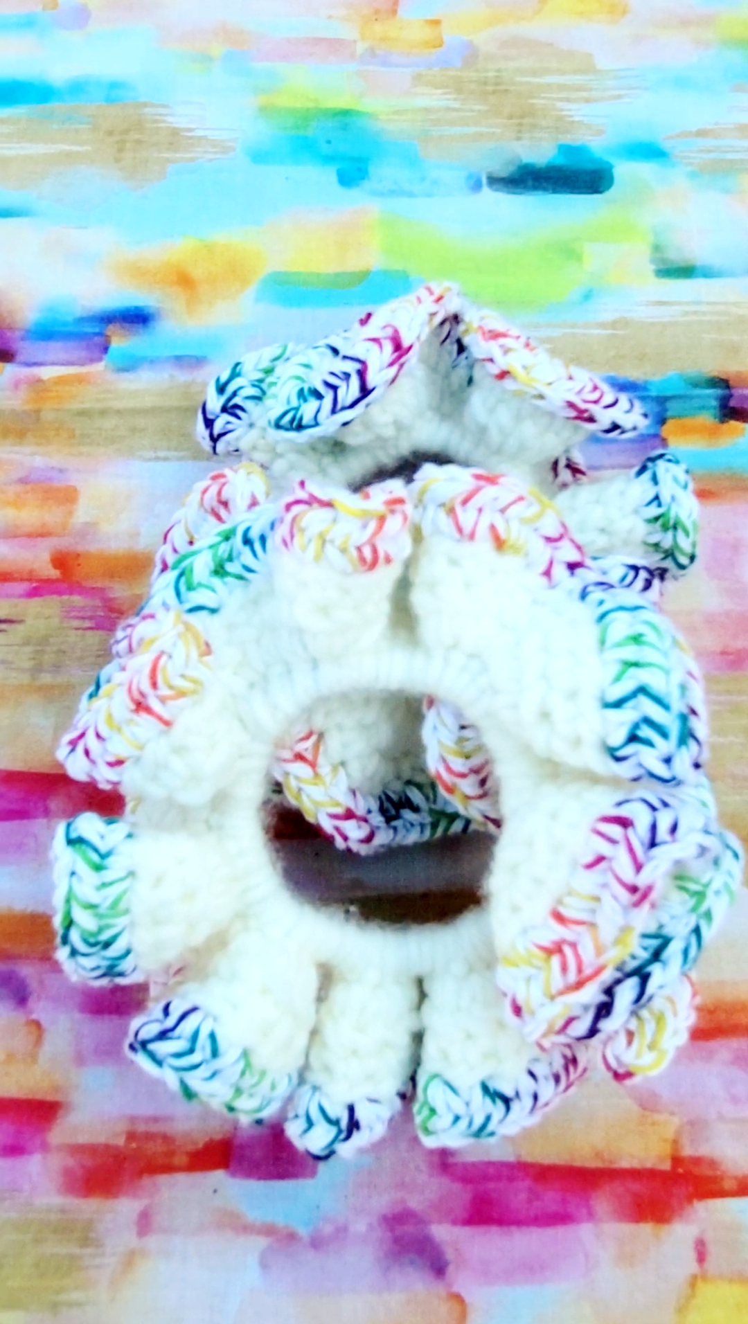 A white crochet hair scrunchie with multicolored braided trim, shown against a colorful painted background.