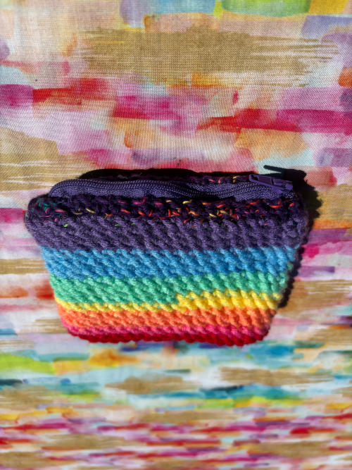 A small handmade crochet card poach with a rainbow stripe pattern and a dark purple zipper, placed on a colorful abstract painted background.