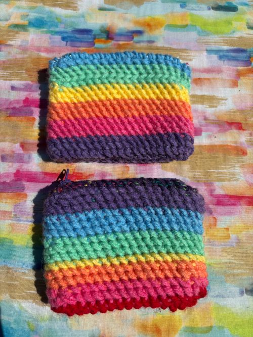 Two handmade rainbow-striped crochet card purses with zipper closures, displayed on a colorful watercolor-textured background.