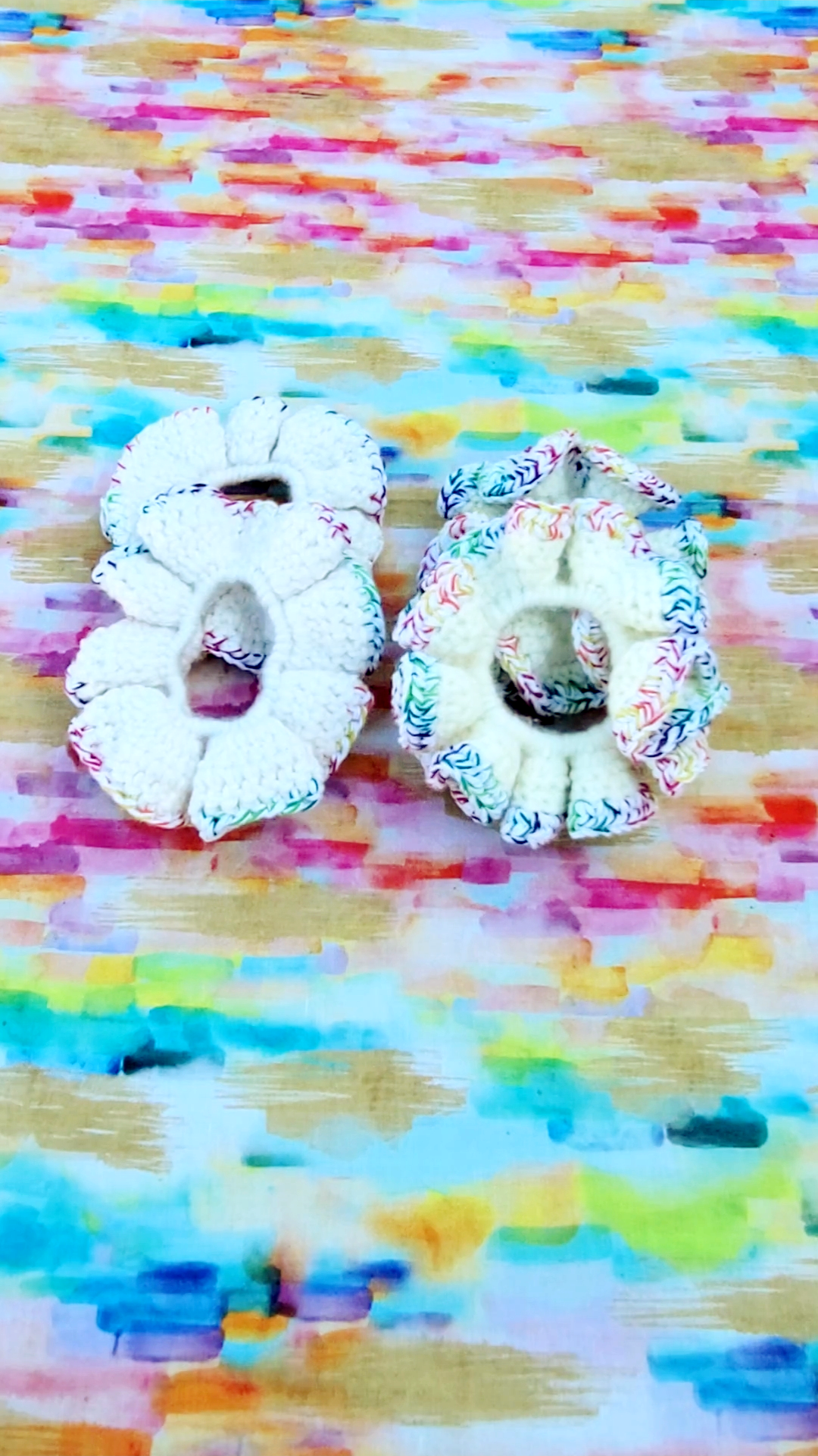Four handmade crochet hair scrunchies, two solid white and two multicolor patterned, displayed on a vibrant, multicolored painted background.