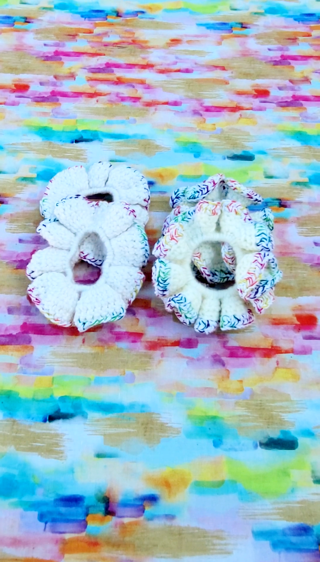 Four crocheted scrunchies with colorful patterned trim, displayed side-by-side on a vibrant, multicolored painted background.