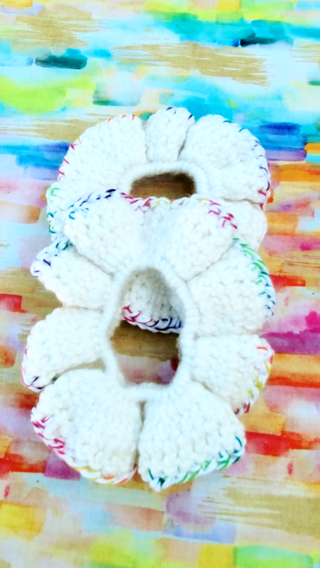 Two crocheted scrunchies with a off white center and trim of white with multiple colors of red, purple, pink, blue, green, yellow and orange.