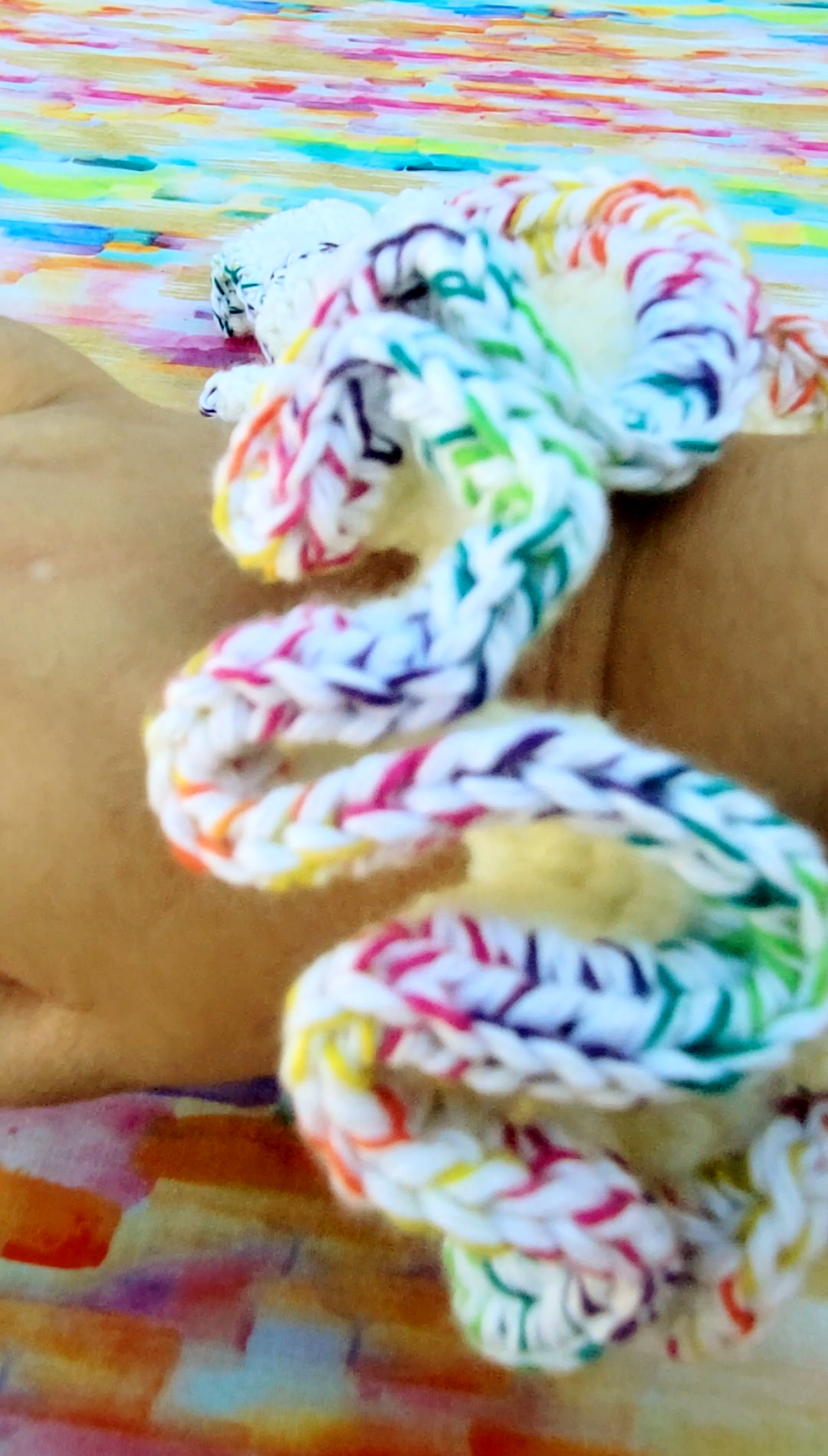 A close-up of a crochet hair scrunchie wrapped around a person's wrist, featuring a multicolor pattern of white braid with pink, blue, green, yellow, and purple accents, set against a colorful painted background.