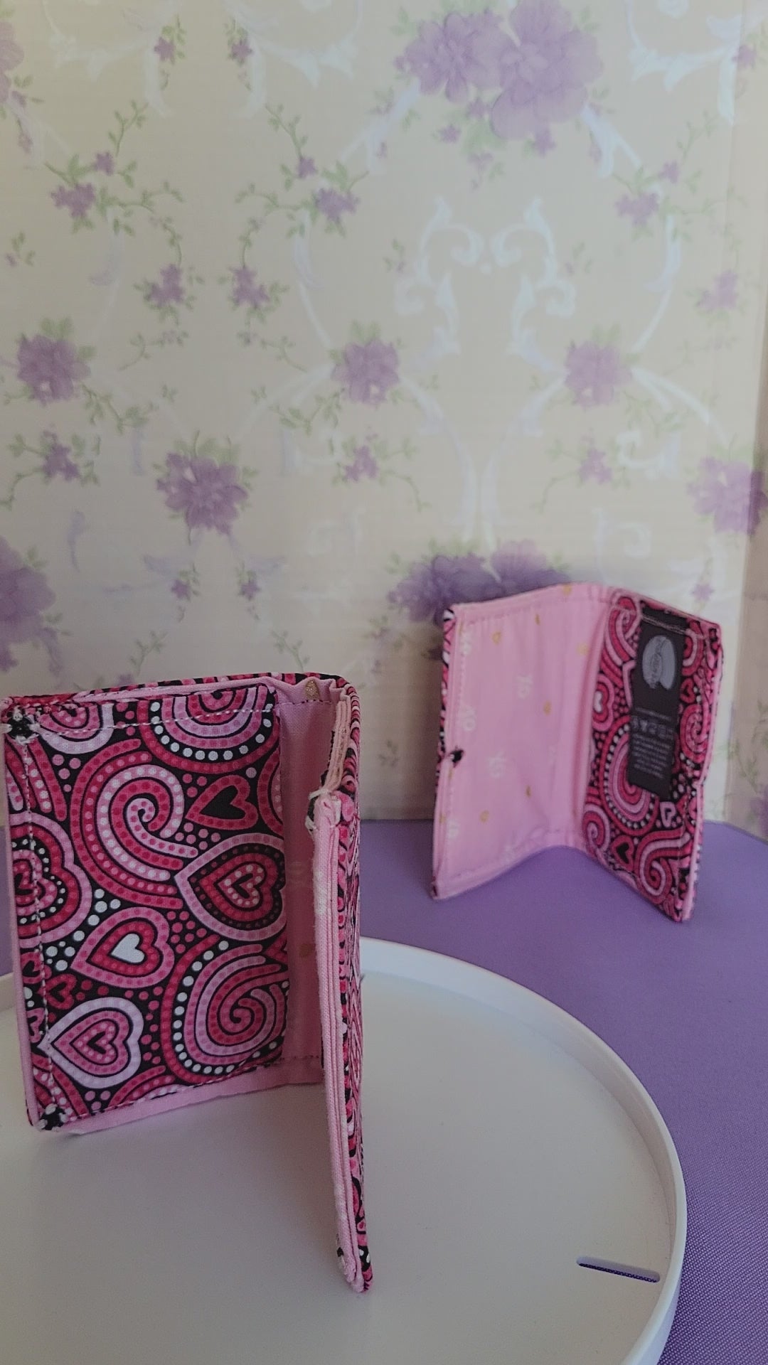 Two Javier wallets with a ripple effect of hearts in a selection of colors pink, white, red and black. They are placed parallel with one on a turning table.
