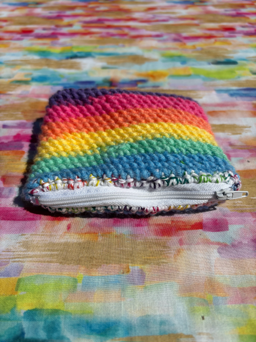 A small handmade zippered card bag made from rainbow-colored yarn, featuring horizontal stripes of pink, orange, yellow, green, blue, and purple, resting on a colorful painted fabric background.